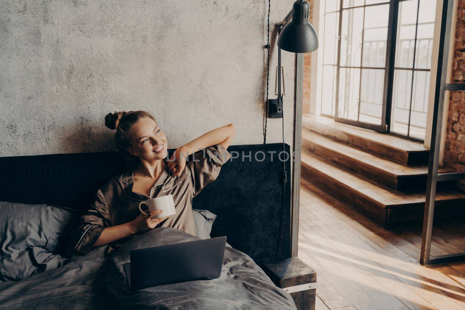 Happy positive young girl in satin pajama lying in bed at home after waking up from night sleep holding her morning cup of coffee and checking new emails on laptop or preparing to start remote work