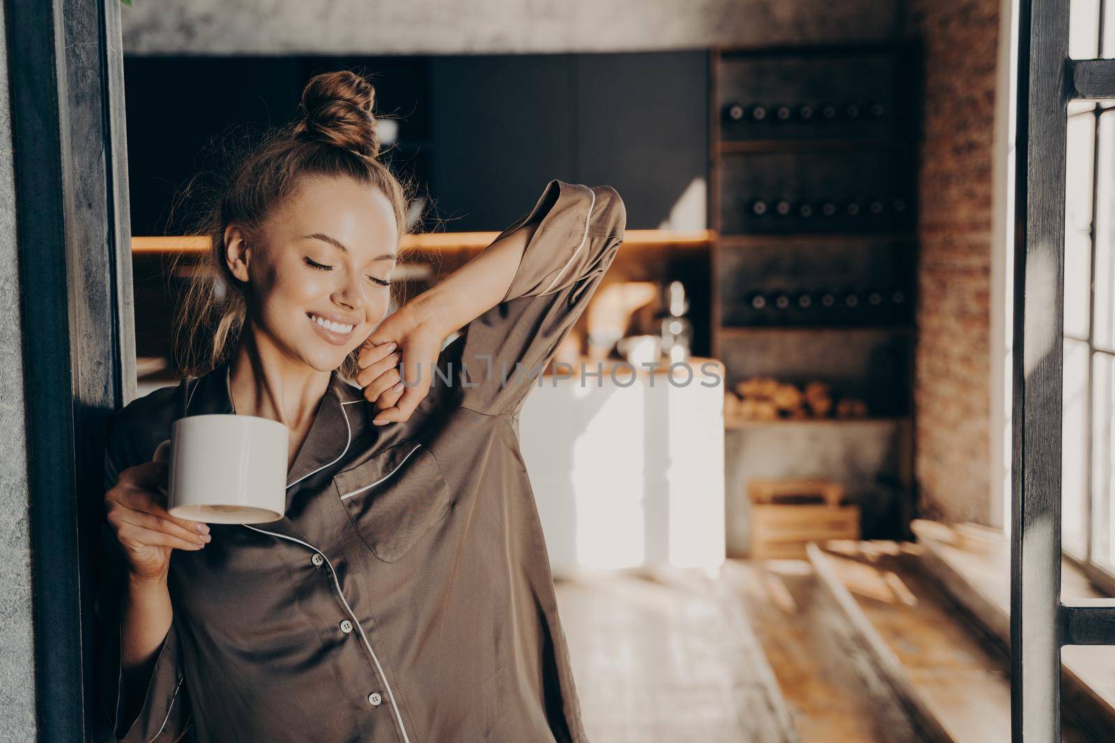 New day. Happy beautiful brunette female stretching with cup of coffee in her hand enjoying morning sunshine while standing in kitchen doorway in satin pajama. People and lifestyle concept