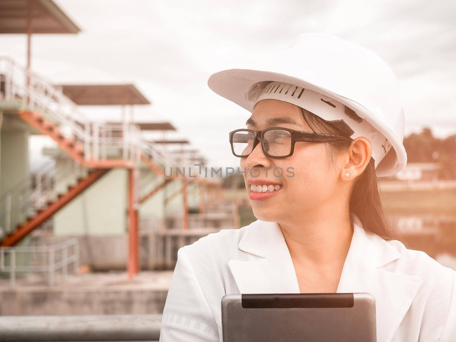 Female engineer in a white helmet talking radio communication (walkie talkie) and holding a digital tablet, stands in construction site of the hydroelectric dam.