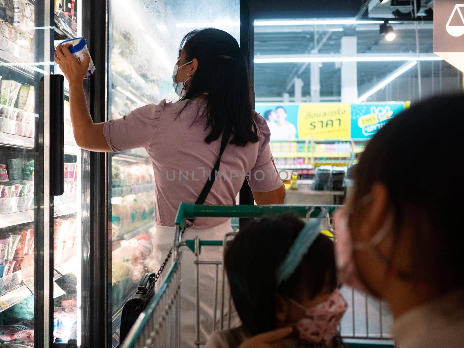 10 July 2021; Mother is choosing fresh dairy products or yogurt in the refrigerator  with her two young daughters sitting in a shopping cart in a supermarket in Chiang Mai. Shopping for healthy.