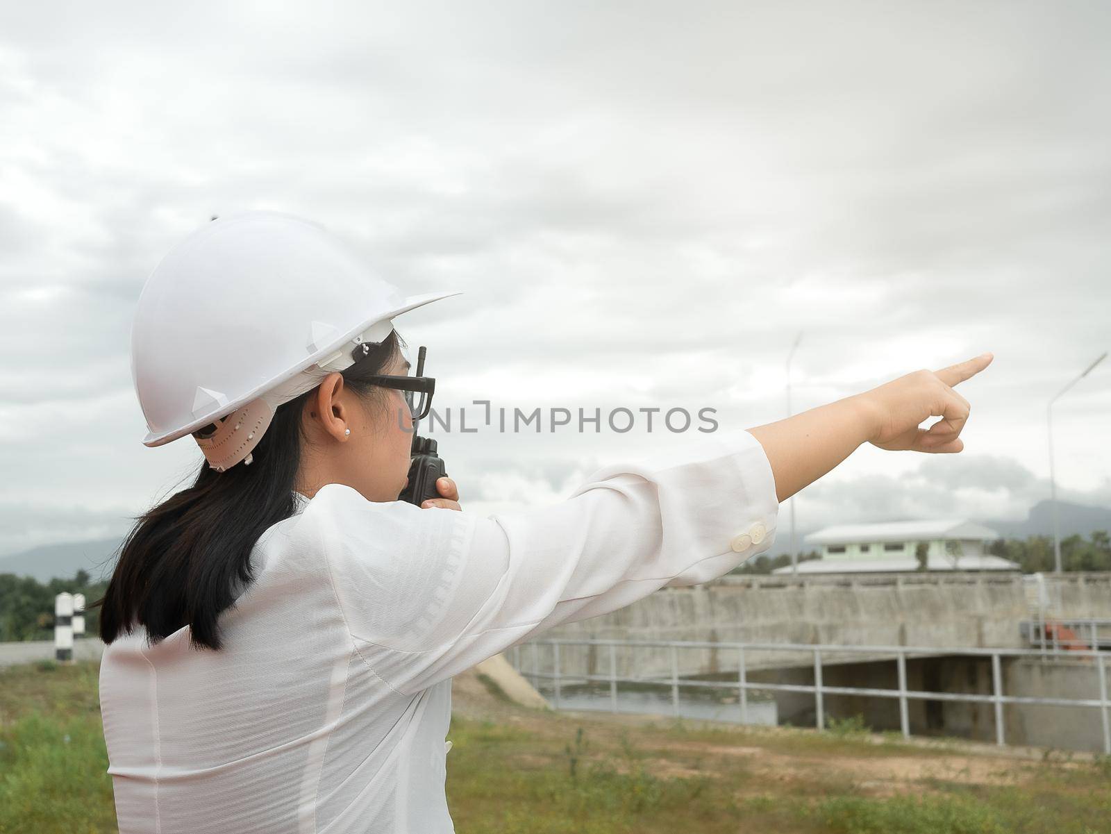 Female engineer in a white helmet using radio communication with construction worker at the dam construction site to generate electricity. by TEERASAK