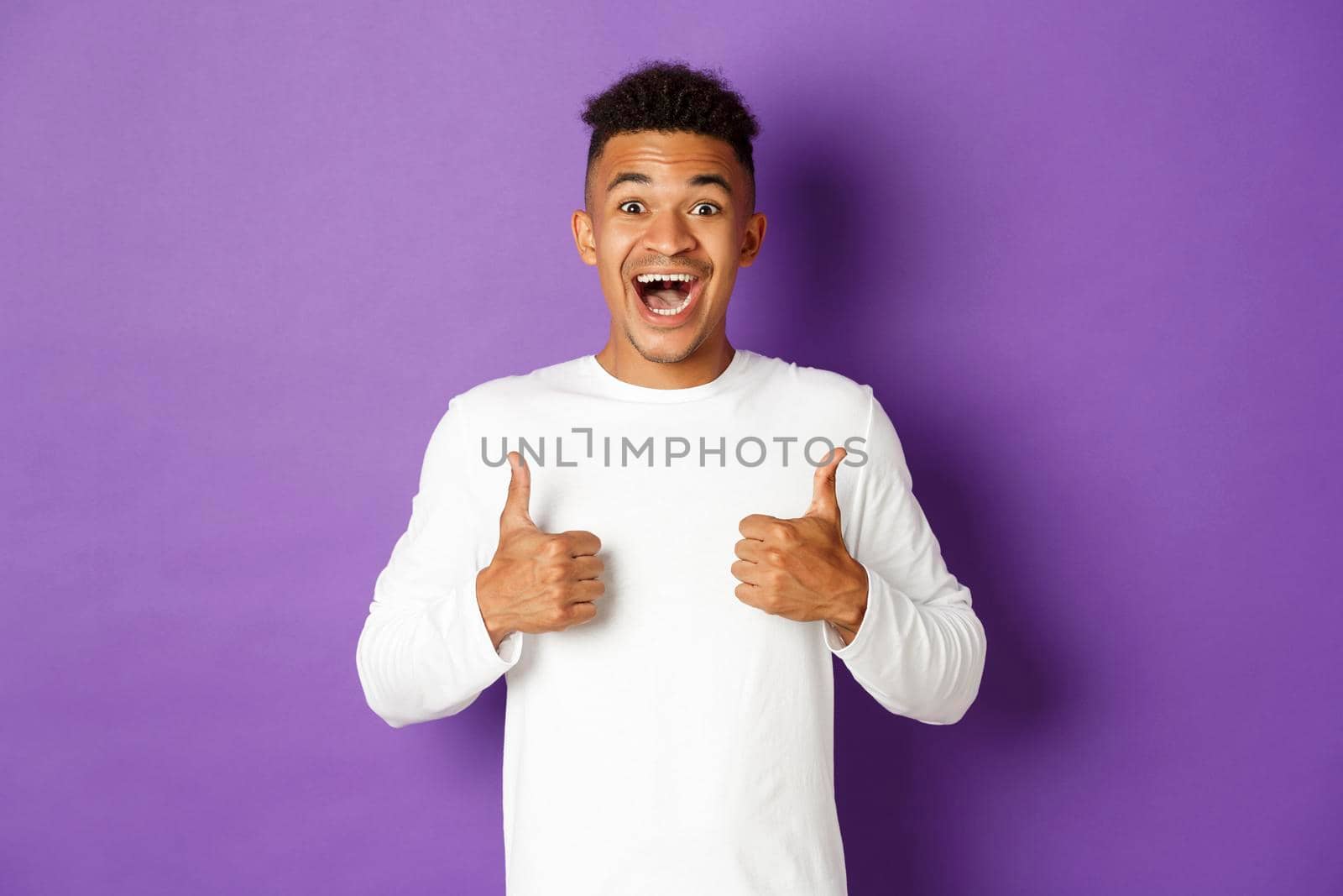 Excited african-american man showing thumbs-up, smiling amazed, like and praise great promo, standing over purple background by Benzoix