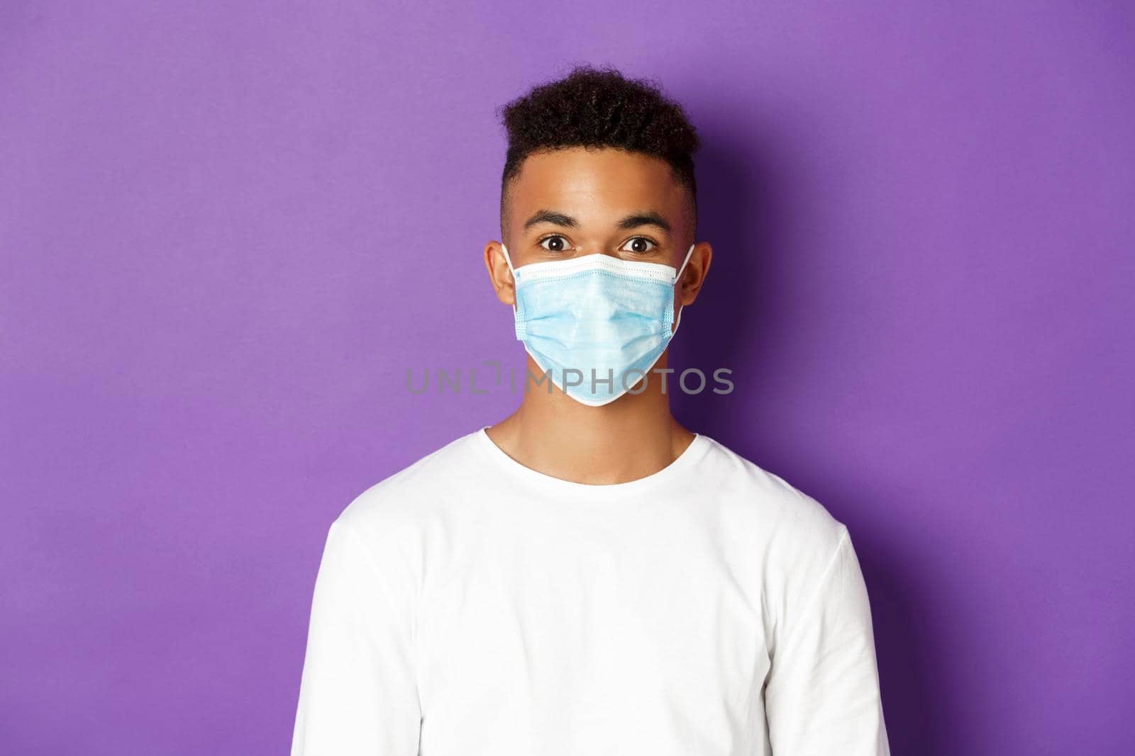 Concept of coronavirus, quarantine and social distancing. Close-up of young african-american guy in white sweatshirt and medical mask, looking at camera, standing over purple background.