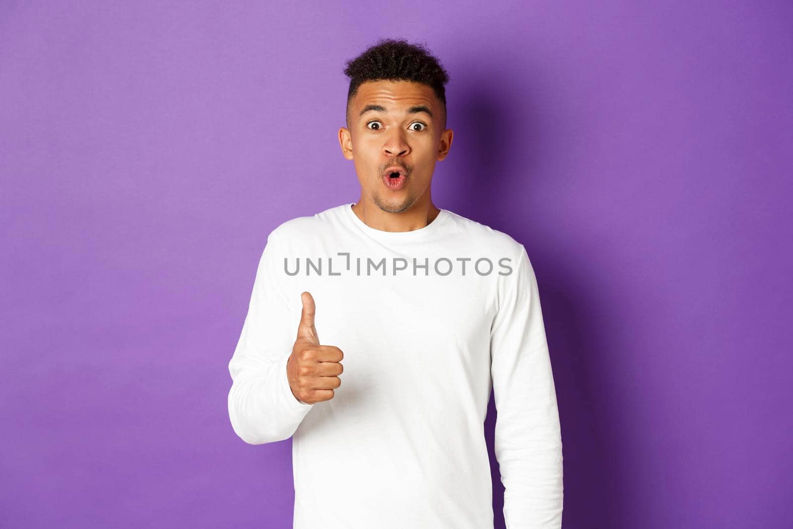 Portrait of amazed african-american man, showing thumbs-up and looking excited, recommend and praise good product, standing over purple background.
