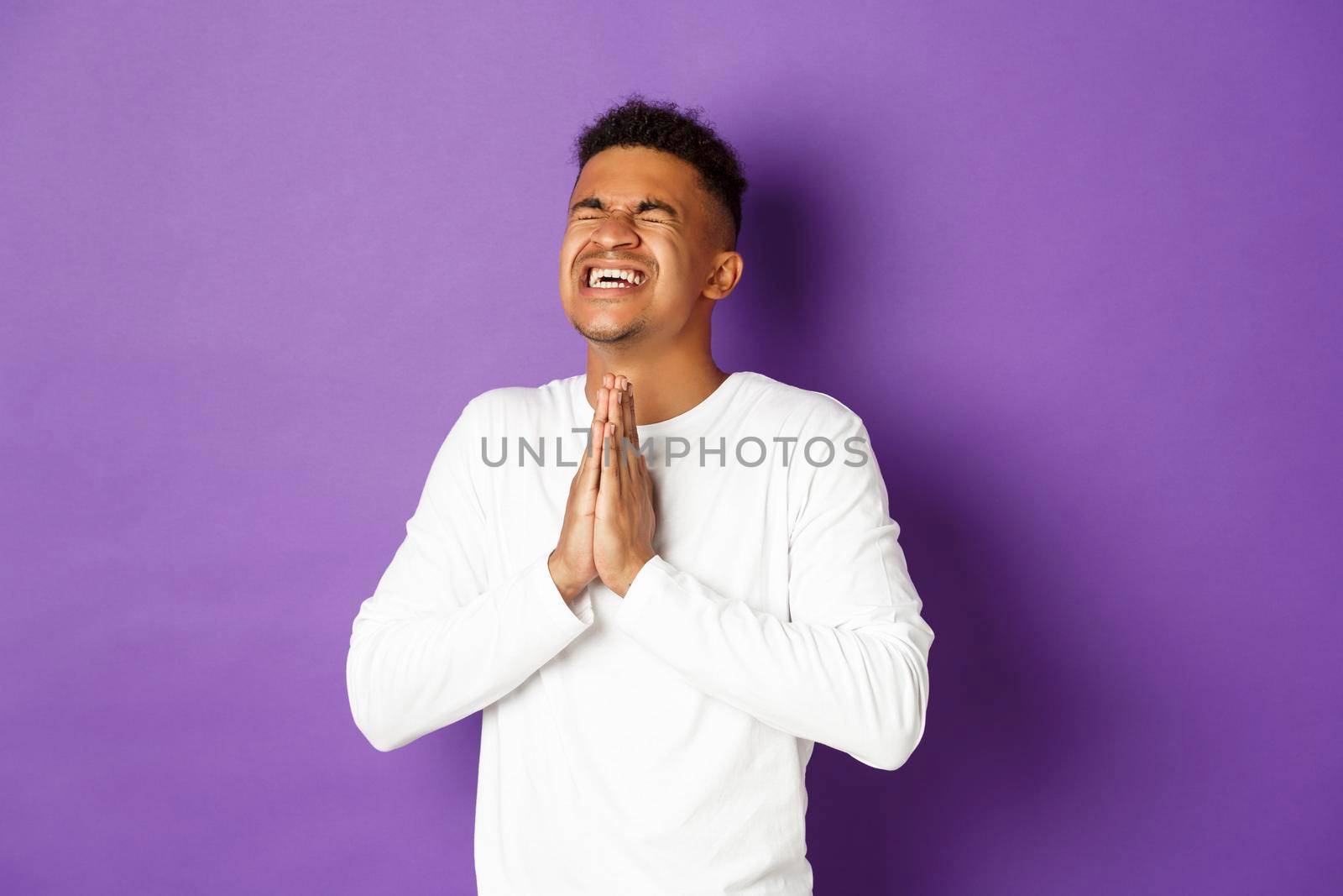 Image of desperate african-american guy pleading to God, holding hands in pray and begging for something, making wish, standing worried over purple background.