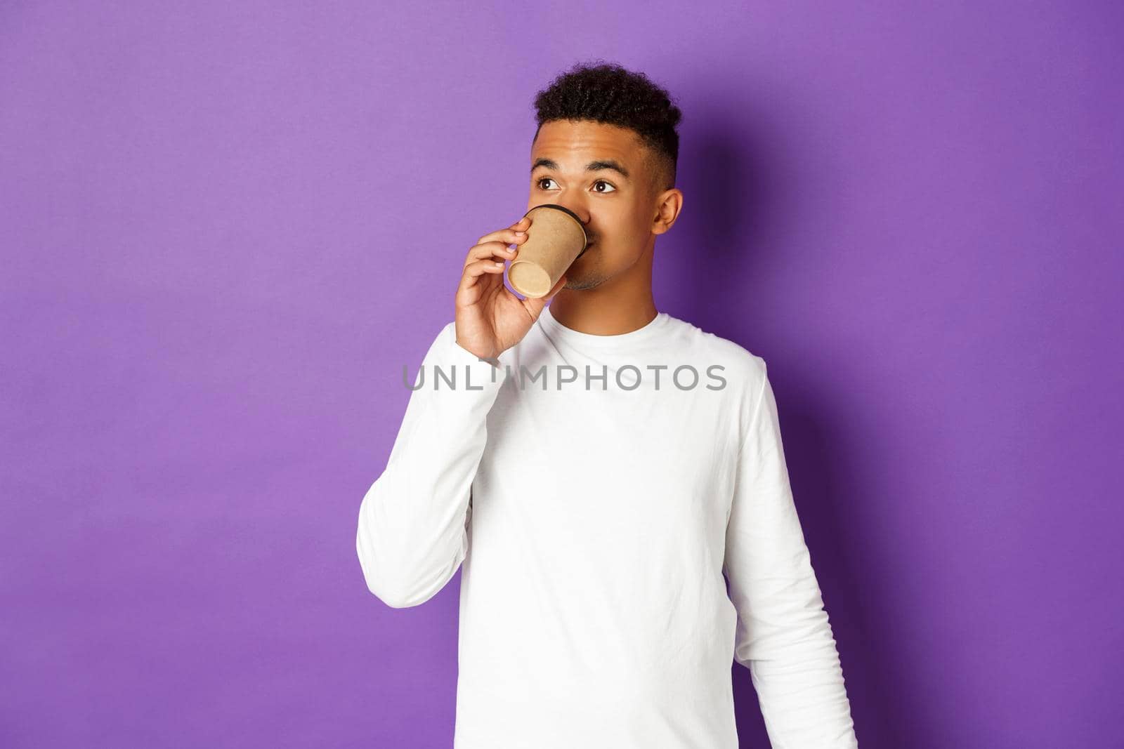 Portrait of young modern african-american guy drinking coffee, looking at upper left corner, standing over purple background.