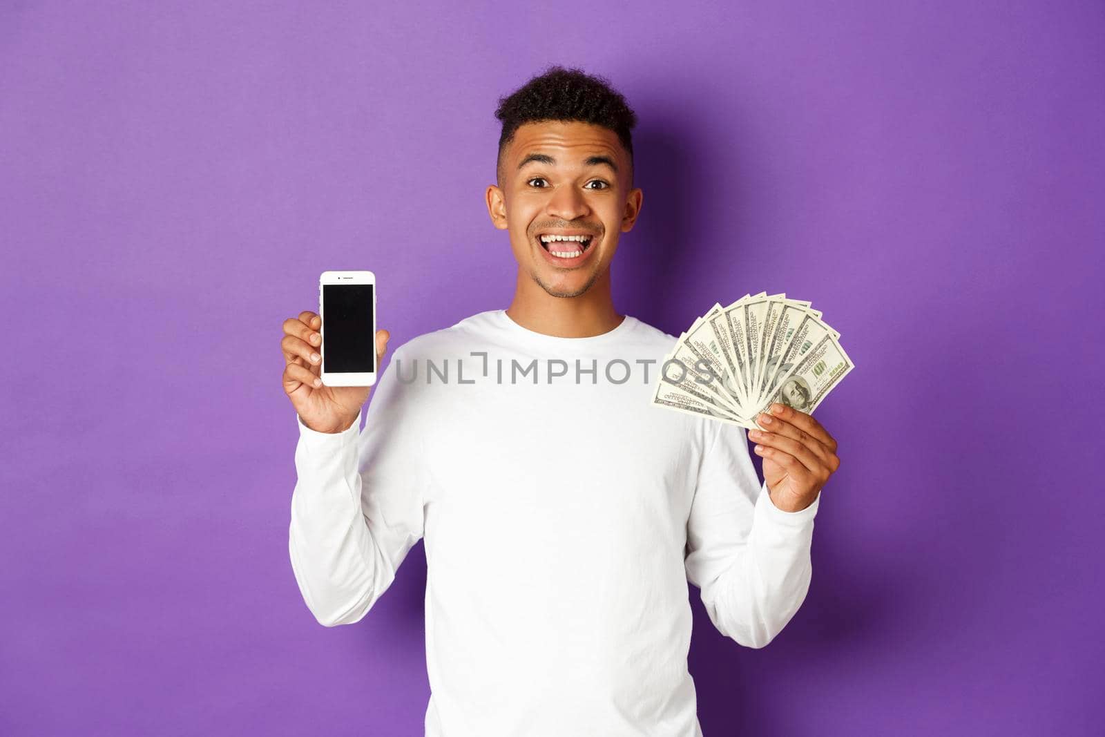 Portrait of cheerful african-american man, showing mobile phone screen and money, standing against purple background.