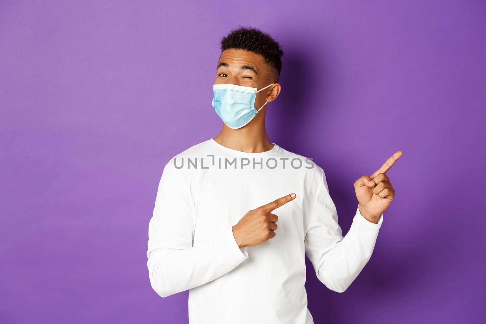Concept of coronavirus, quarantine and lifestyle. Cheerful african-american guy in medical mask, winking and pointing at upper right corner, showing logo, standing over purple background.