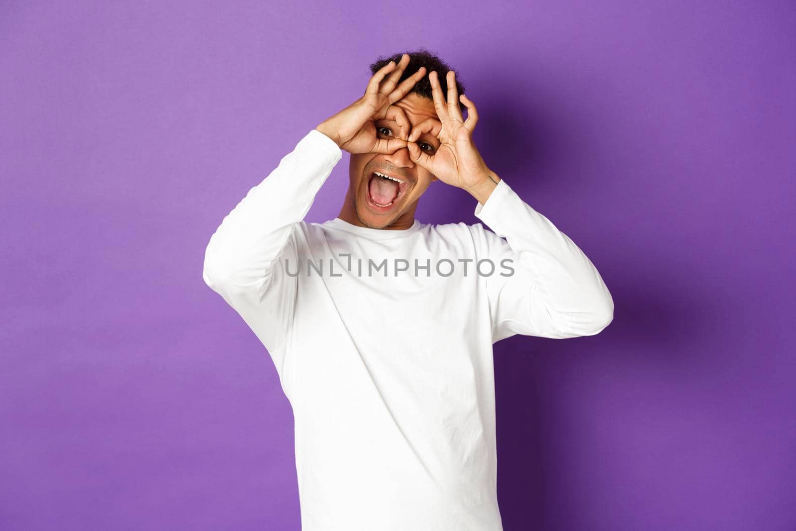 Image of funny and happy african-american man in white sweatshirt, grimacing and making faces, standing over purple background by Benzoix