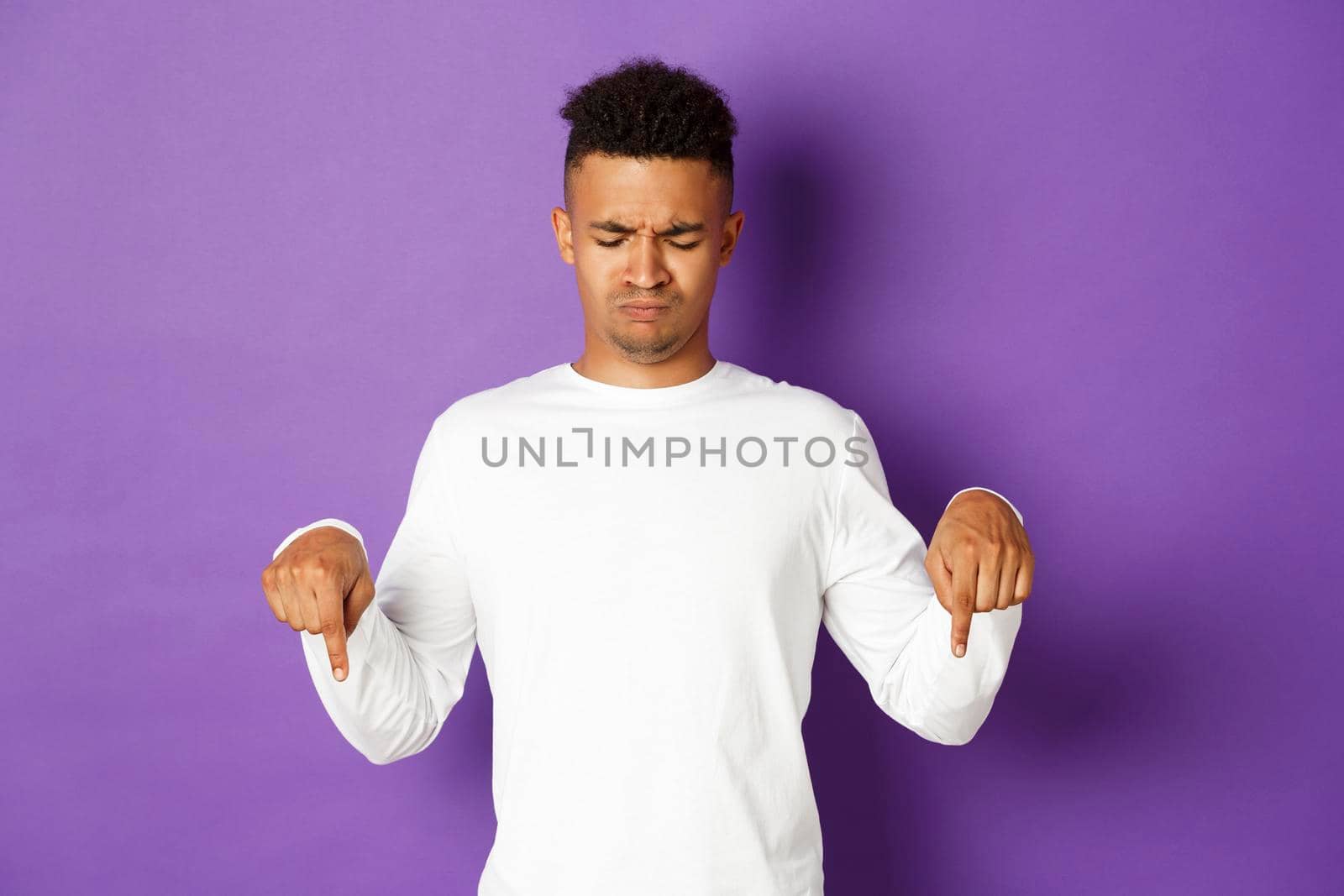 Portrait of skeptical and disappointed african-american male model in white sweatshirt, frowning and pouting upset, pointing and looking down, standing over purple background.