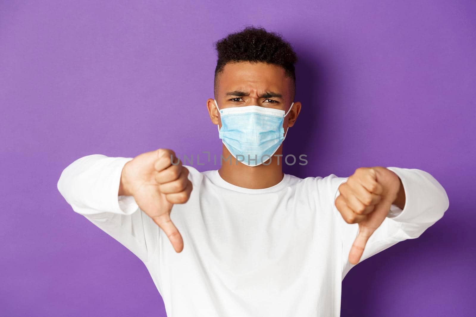 Concept of coronavirus, quarantine and social distancing. Close-up of young african-american man in medical mask showing thumbs-down, dislike something bad, standing over purple background.