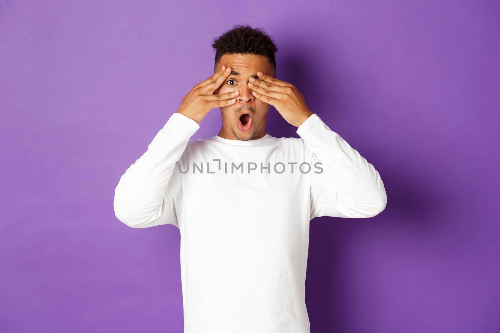 Portrait of curious african-american man, waiting for surprise, close eyes with hands but peeking through fingers at something interesting, standing over purple background by Benzoix