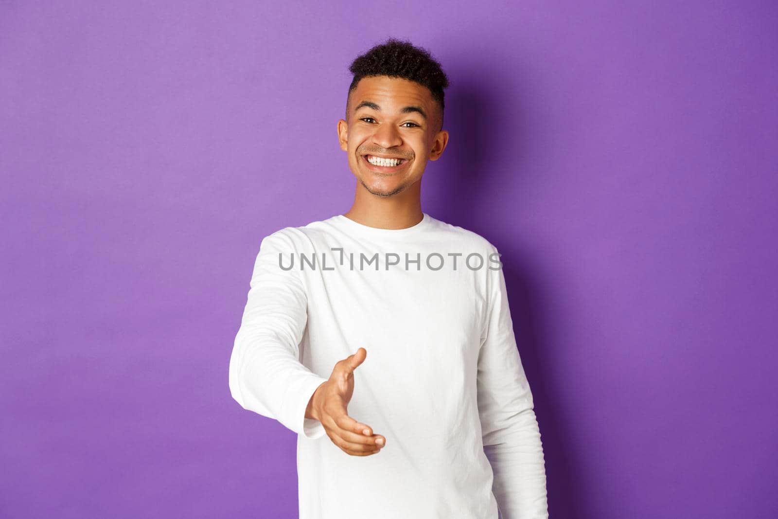 Image of handsome young african-american man, smiling friendly while extending hand for handshake, greeting someone, standing over purple background.