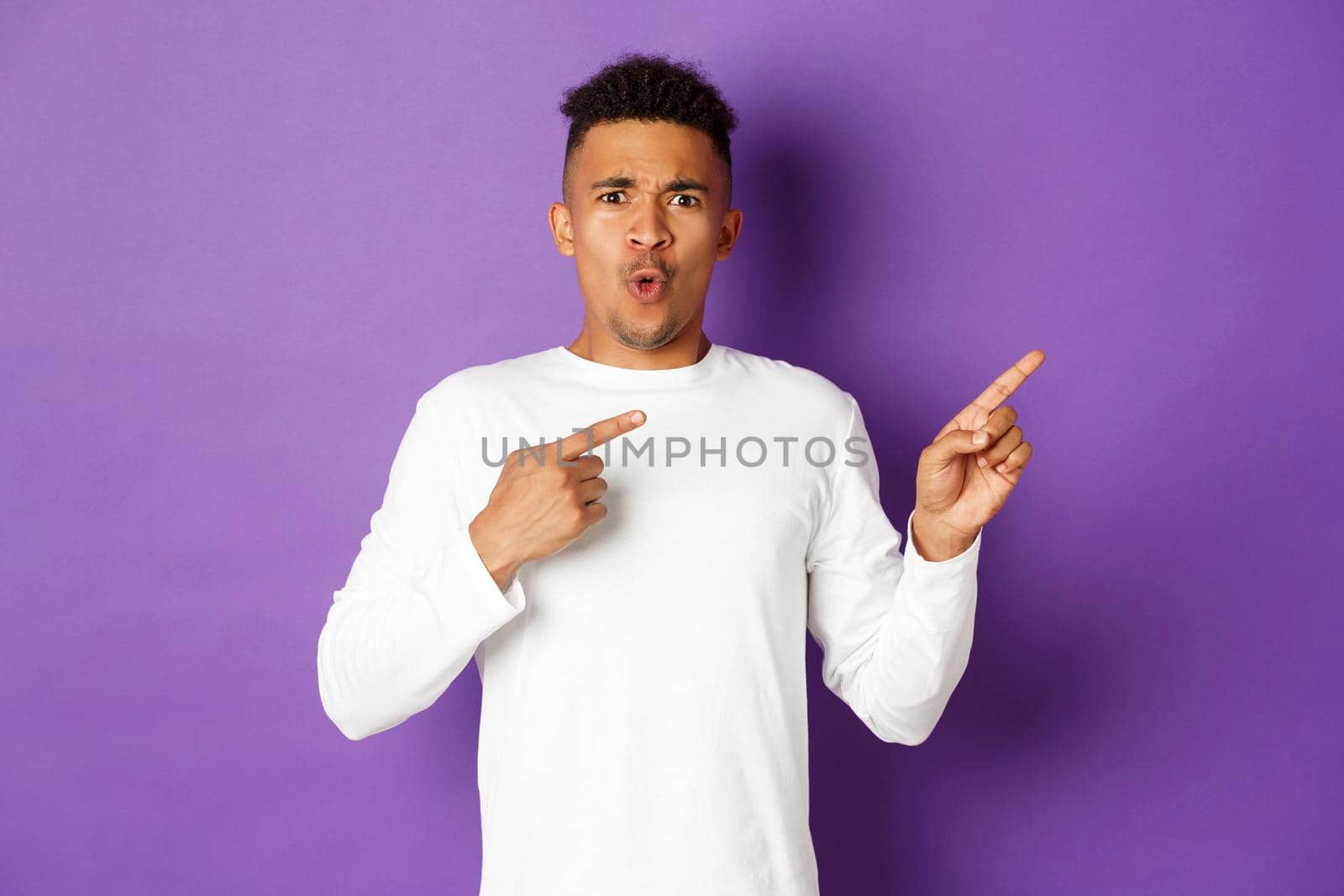 Portrait of confused and shocked african-american guy in white sweatshirt, pointing fingers right and frowning, showing something strange, standing over purple background by Benzoix