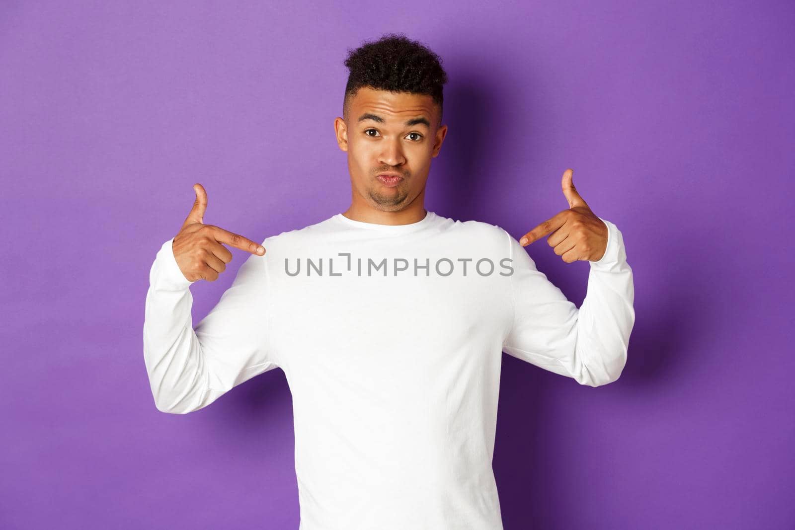 Image of silly and cute african-american guy in white sweatshirt, pointing fingers at your logo on the center, standing over purple background.