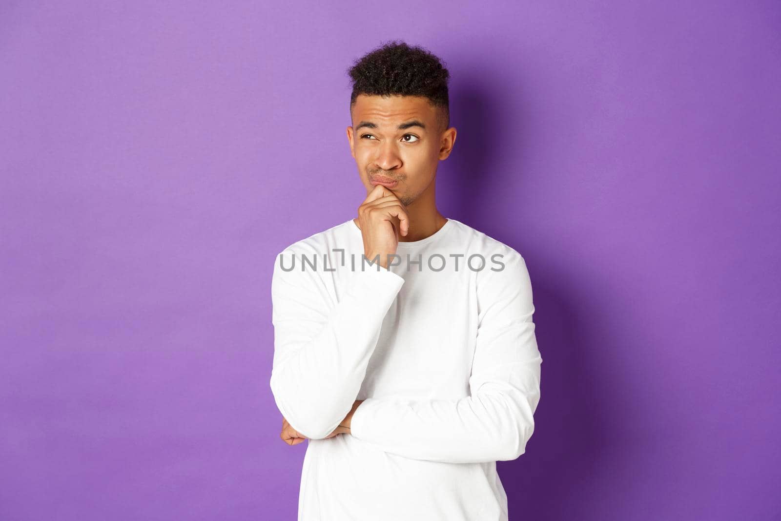 Portrait of young african-american male student thinking, looking at upper left corner thoughtful, making choice, standing over purple background with copy space.