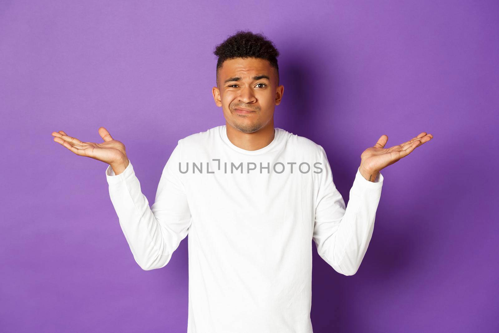 Portrait of indecisive african-american guy in white sweatshirt, raise hands sideways and shrugging clueless, standing confused against purple background.