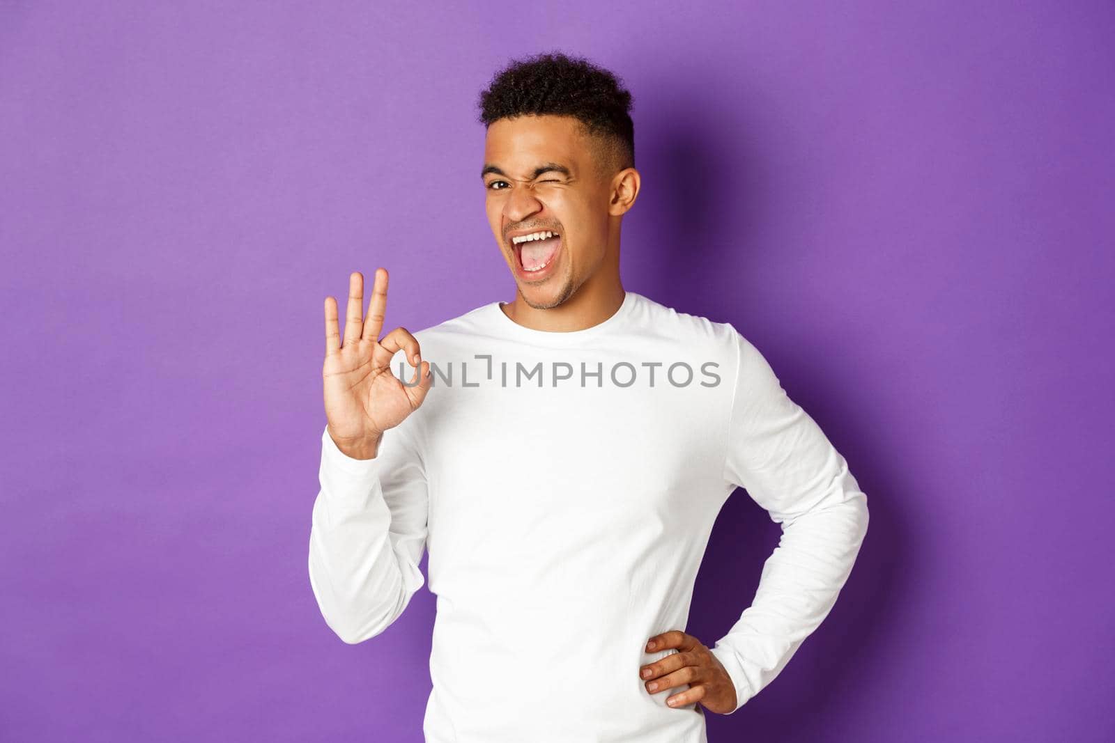 Image of handsome african-american guy, winking cheeky and smiling, showing okay sign, approve something, being satisfied, standing over purple background.