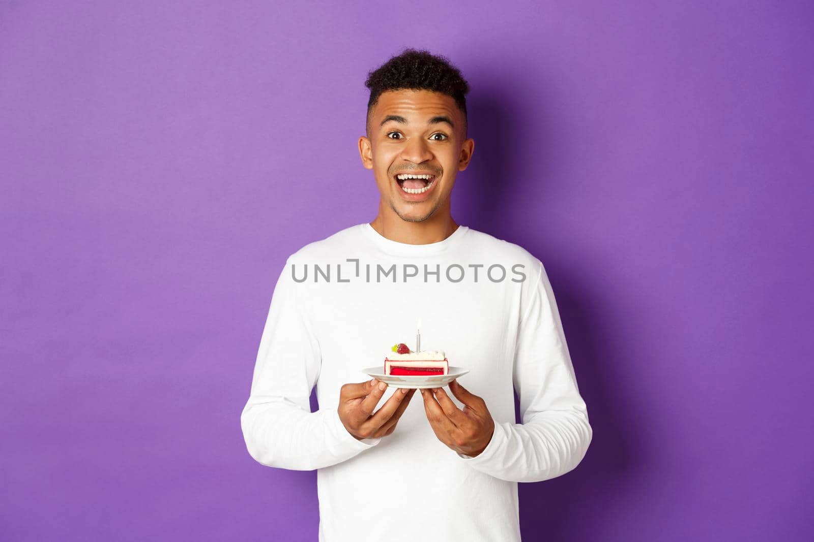 Excited african-american man smiling, holding birthday cake and making wish, celebrating b-day, standing over purple background.