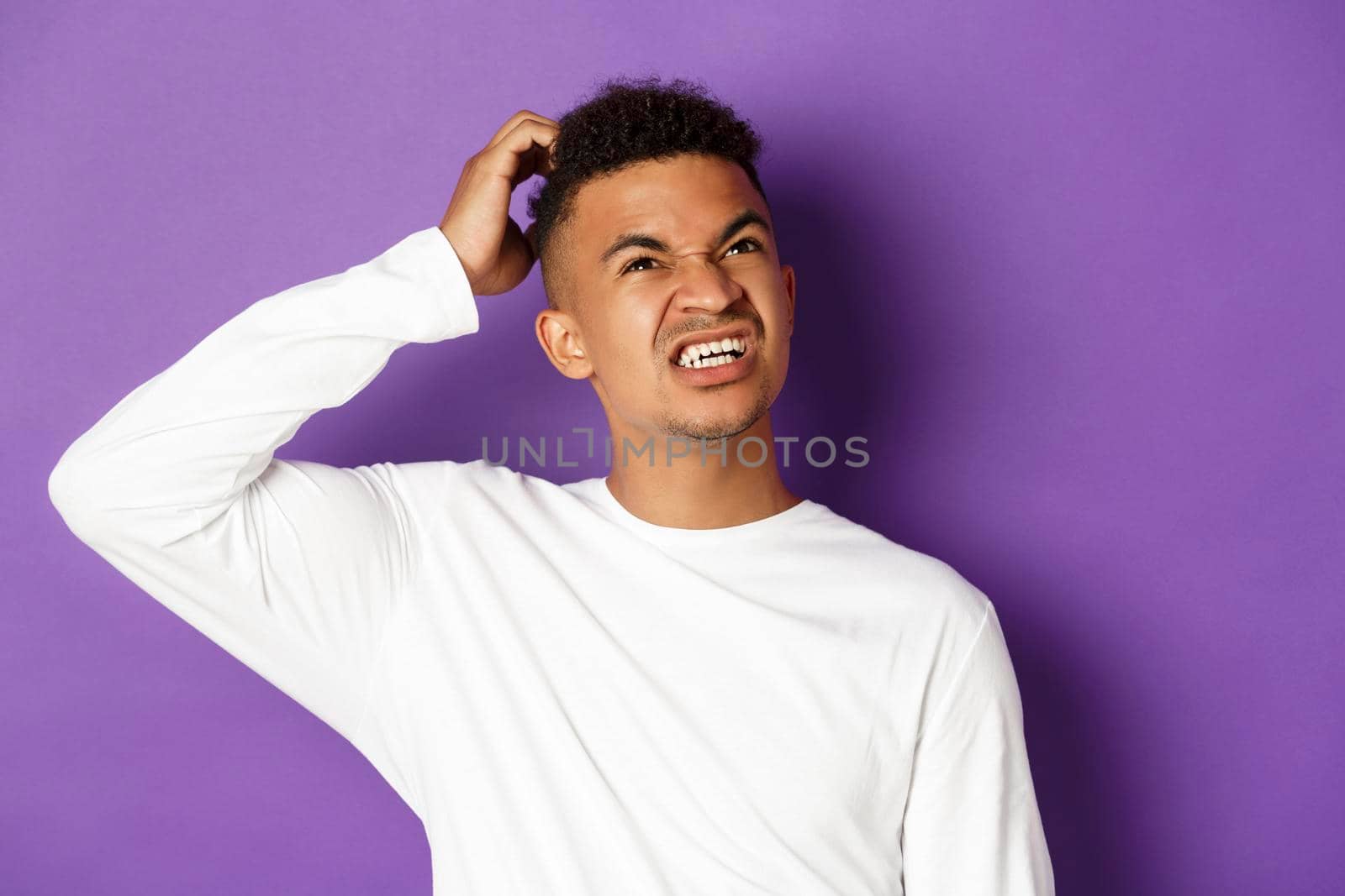 Close-up of troubled african-american man thinking, scratch head and looking up confused, standing in white sweatshirt over purple background.