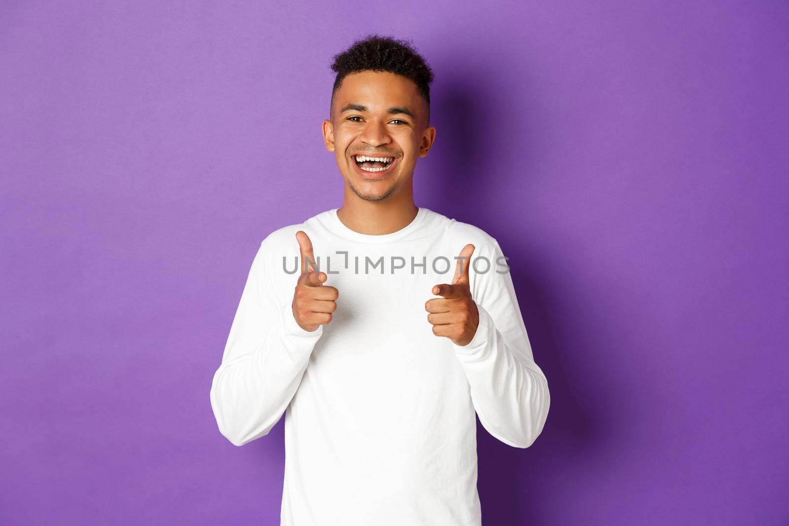 Image of cheerful african-american young guy, pointing fingers at camera and smiling, praise good choice, congratulate with something, standing over purple background.