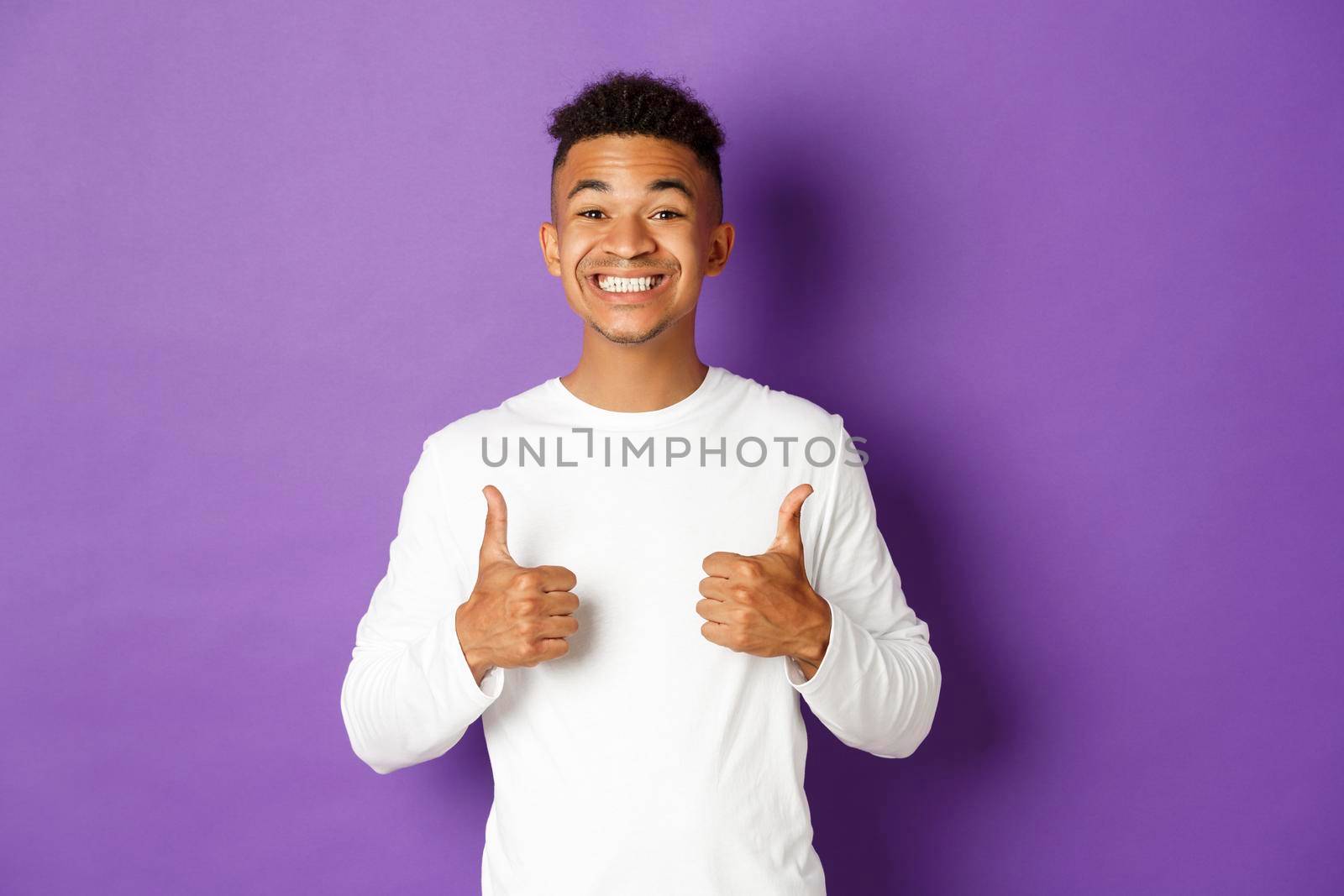 Handsome cheerful man in white sweatshirt, showing thumbs-up in approval and smiling, like and praise something good, standing over purple background.