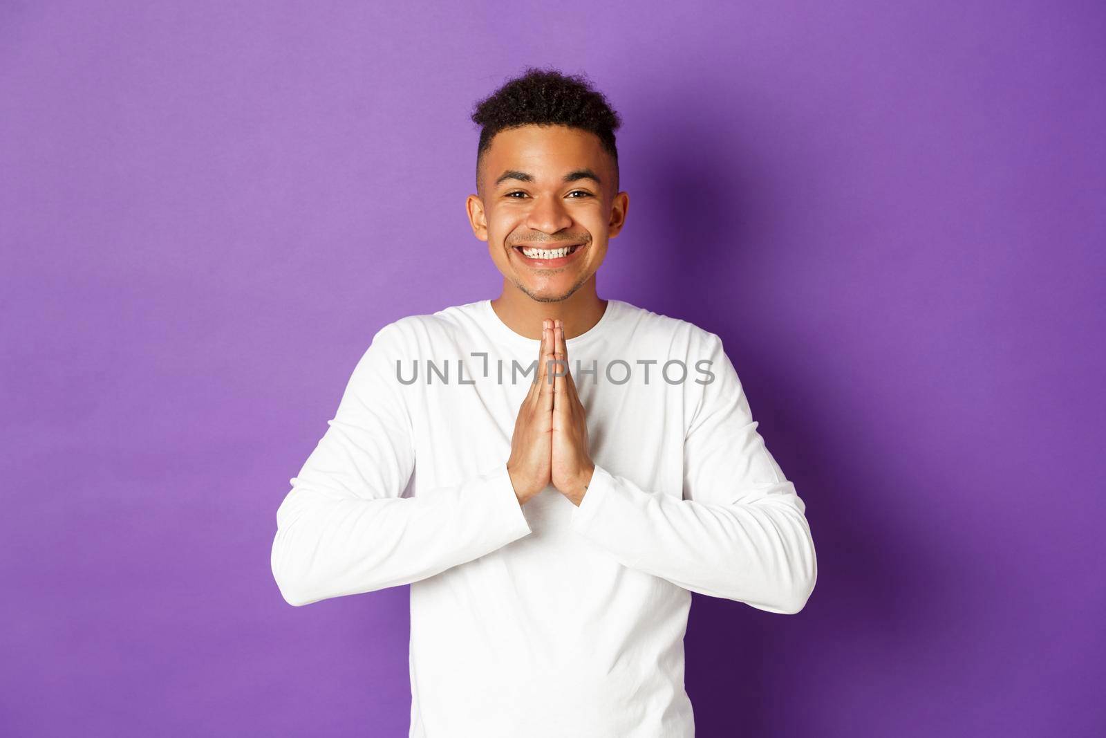 Image of smiling handsome african-american man, thanking for help, holding hands pressed together in pray gesture, looking grateful and happy, standing over purple background by Benzoix