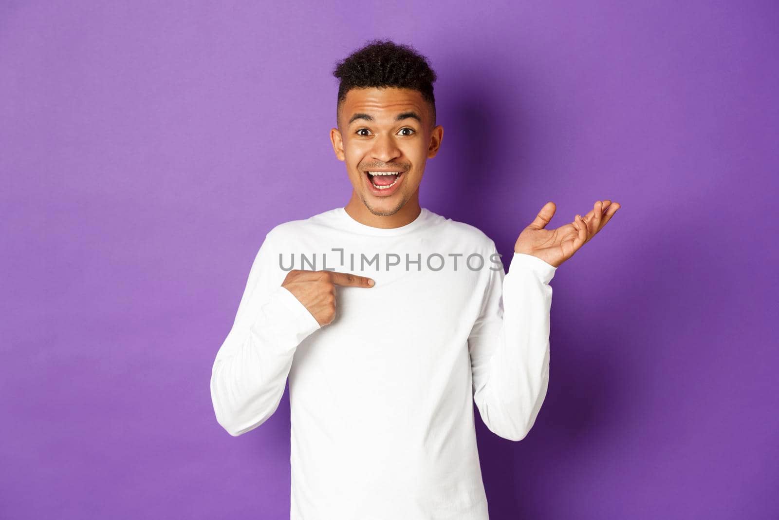 Image of surprised african-american man in white sweatshirt, pointing at himself and raising hand amazed, smiling happy, standing over purple background.