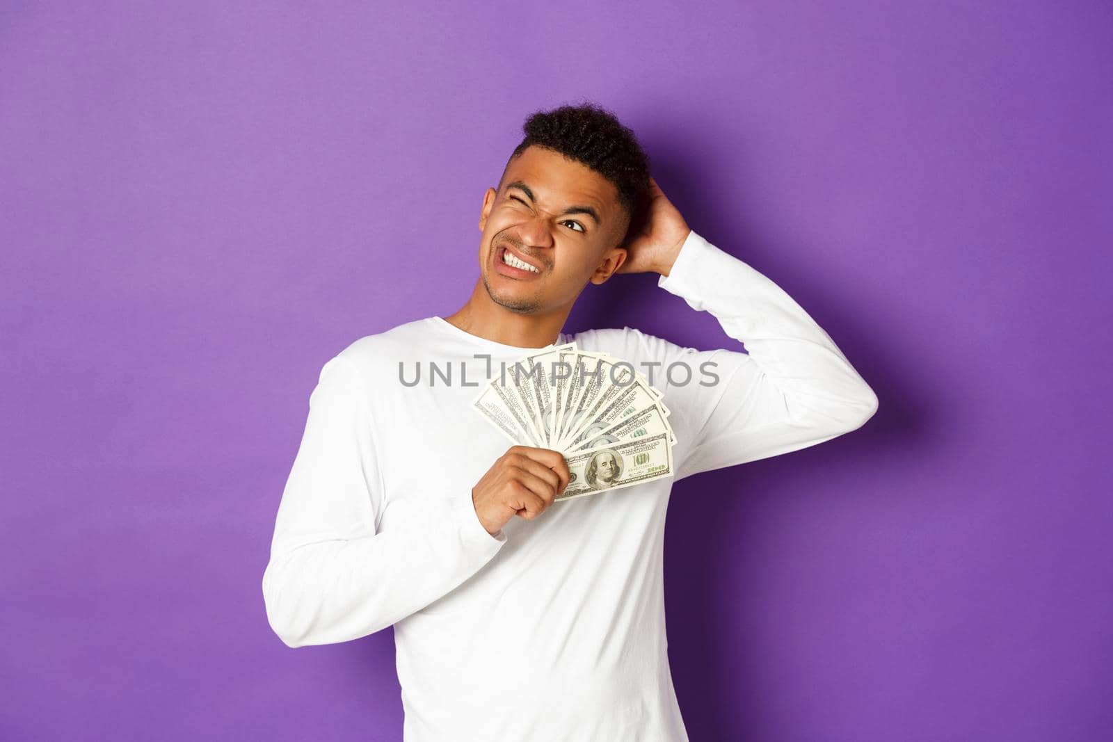 Image of confused african-american man, holding cash and looking at upper left corner puzzled, thinking what to buy, standing over purple background by Benzoix