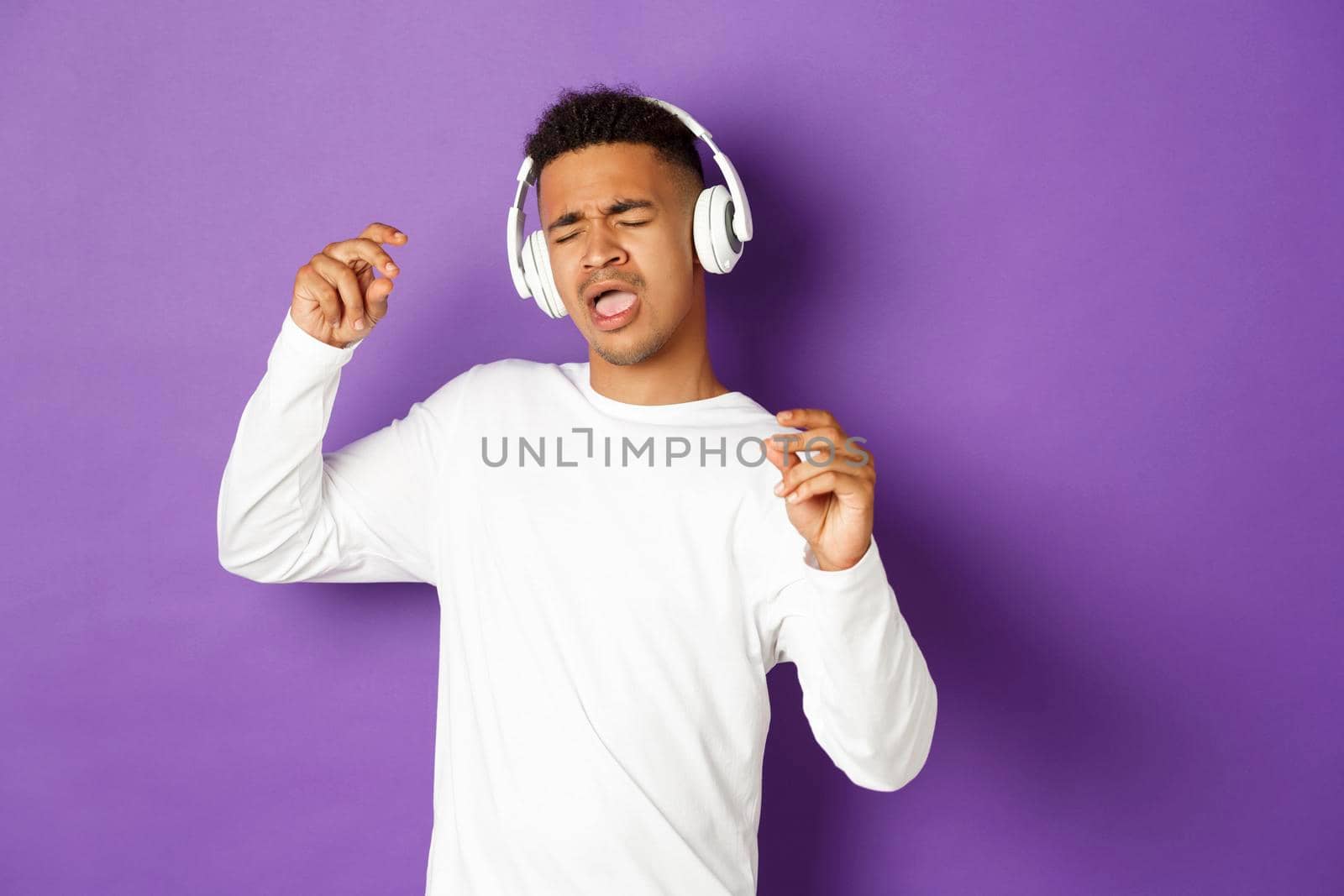 Image of carefree african-american guy dancing, listening music in wireless headphones and singing along, standing over purple background.