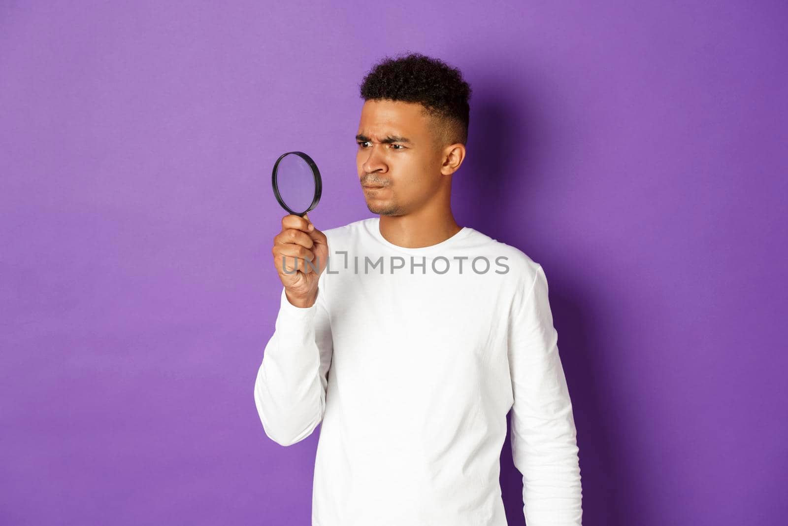 Image of confused african-american man, looking through magnifying glass at something strange, standing over purple background by Benzoix