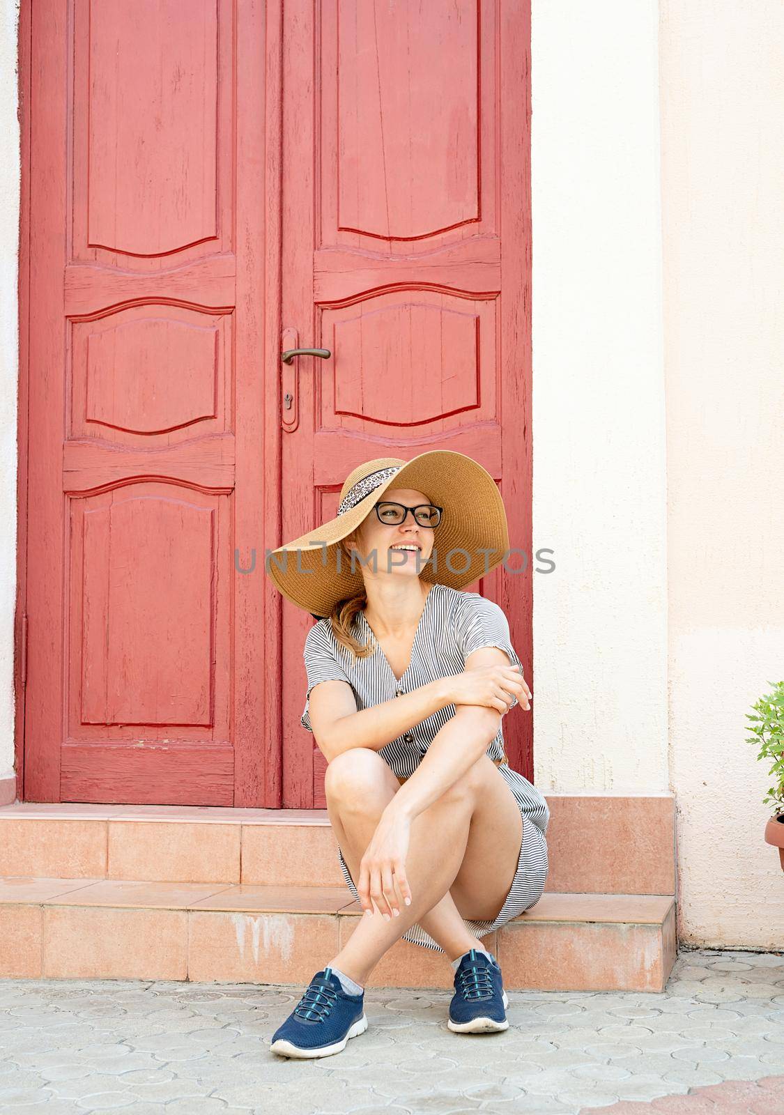 architecture details. Young beautiful woman in summer clothes and glasses sitting on strairs next to the bright red door outdoors