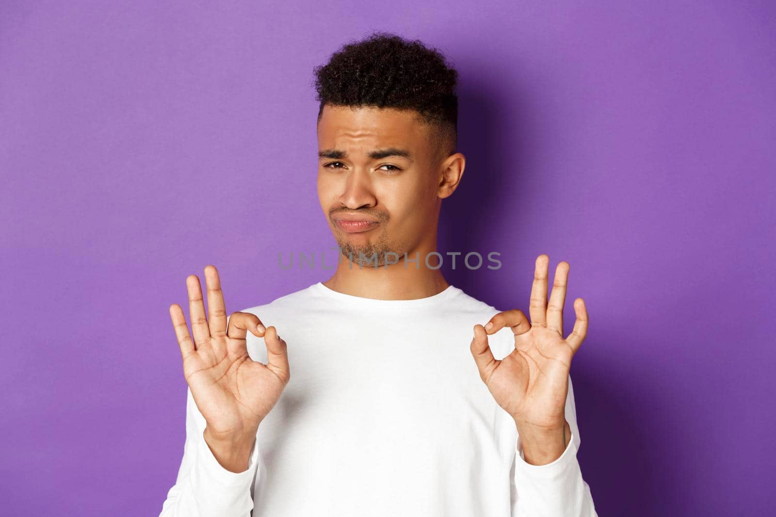Image of pleased african-american man in white sweatshirt, praise something good, showing okay signs and nod in approval, standing over purple background.