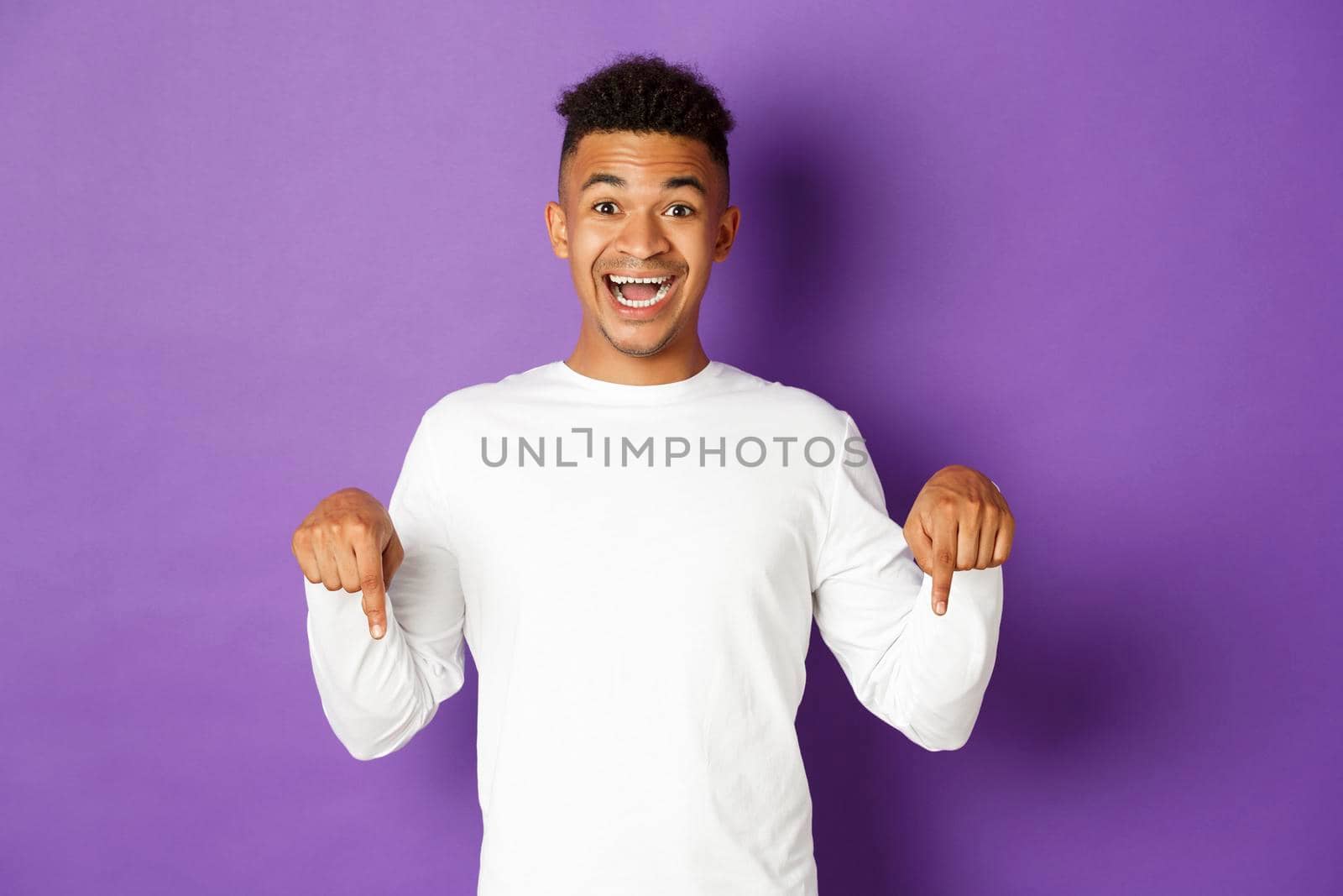 Portrait of handsome african-american guy in white sweatshirt, pointing fingers down and smiling happy, showing promo, looking amazed, standing against purple background.