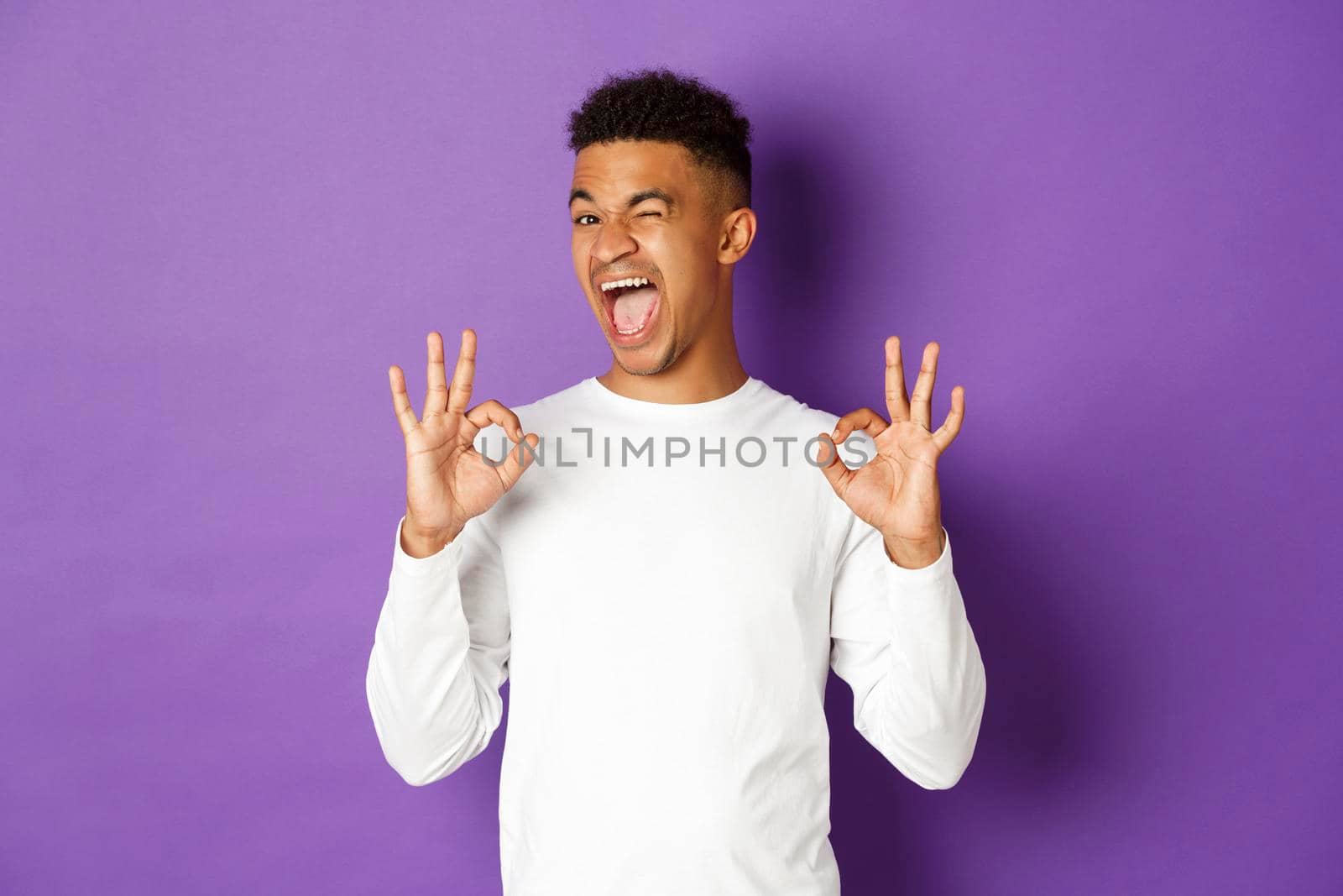 Image of happy and pleased african-american guy, winking and smiling, showing okay signs, recommend good promo offer, like and approve something, standing over purple background by Benzoix