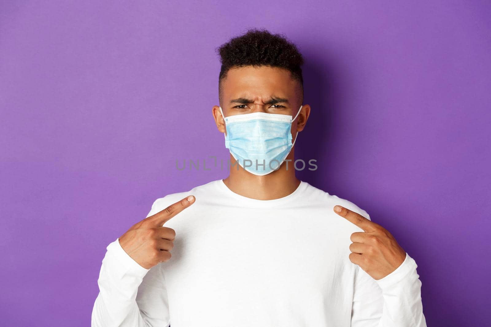 Concept of coronavirus, quarantine and social distancing. Close-up of annoyed african-american man pointing at medical mask, frowning bothered, complaining, standing over purple background.