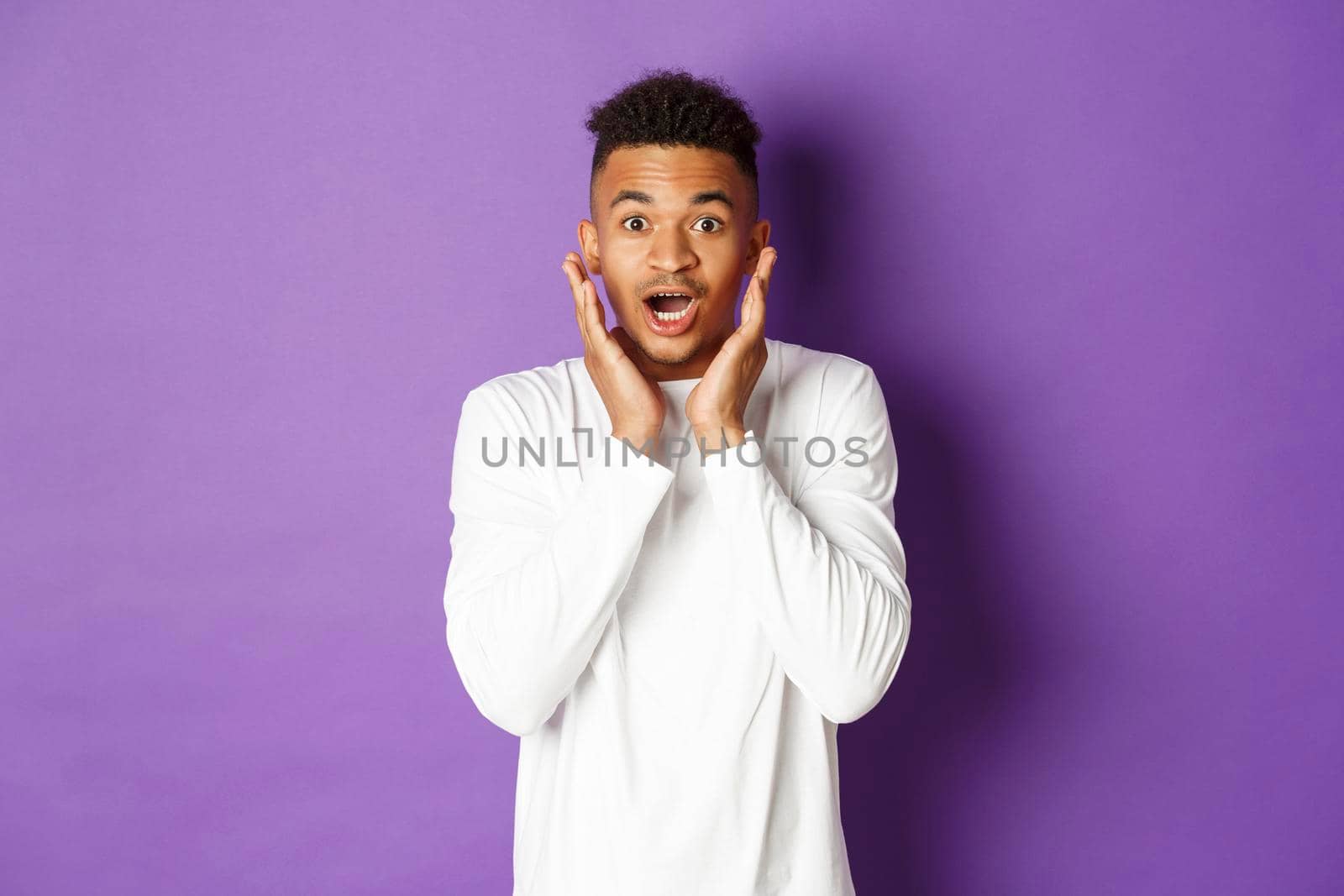 Portrait of handsome african-american male model in white sweatshirt, looking amazed and impressed, gasping wondered, standing over purple background.
