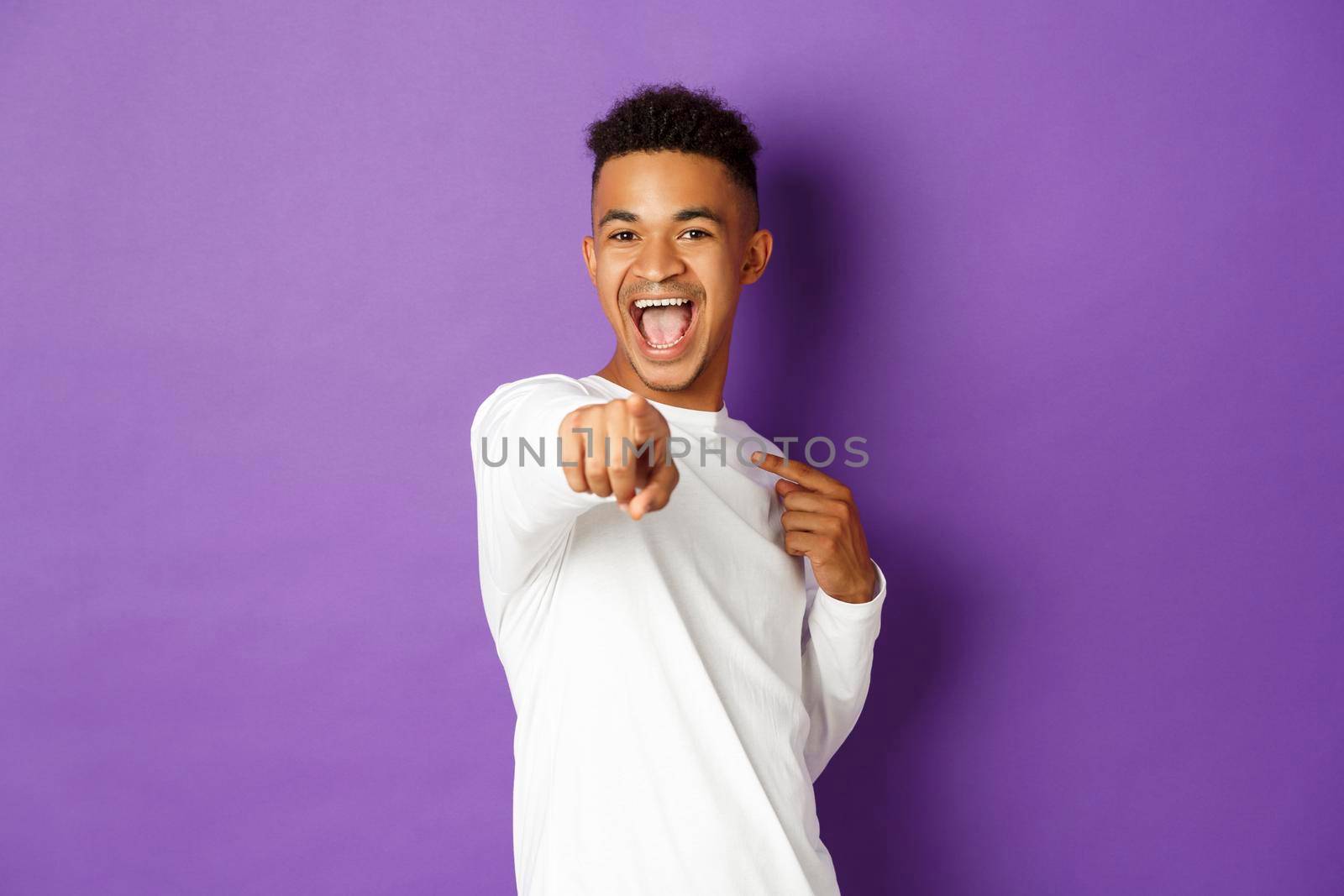 Portrait of cheerful african-american man in white sweatshirt, choosing you, pointing finger at camera to congratulate with something, standing over purple background.