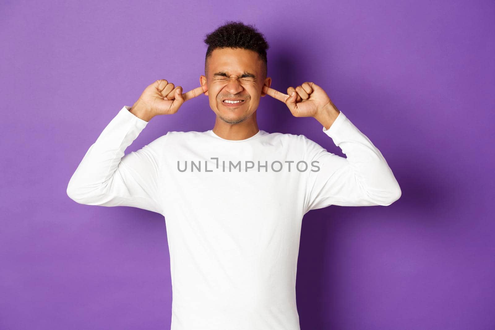 Image of annoyed and disturbed african-american man, complaining on loud noise, grimacing and shut ears with fingers, ignore sound, standing over purple background.