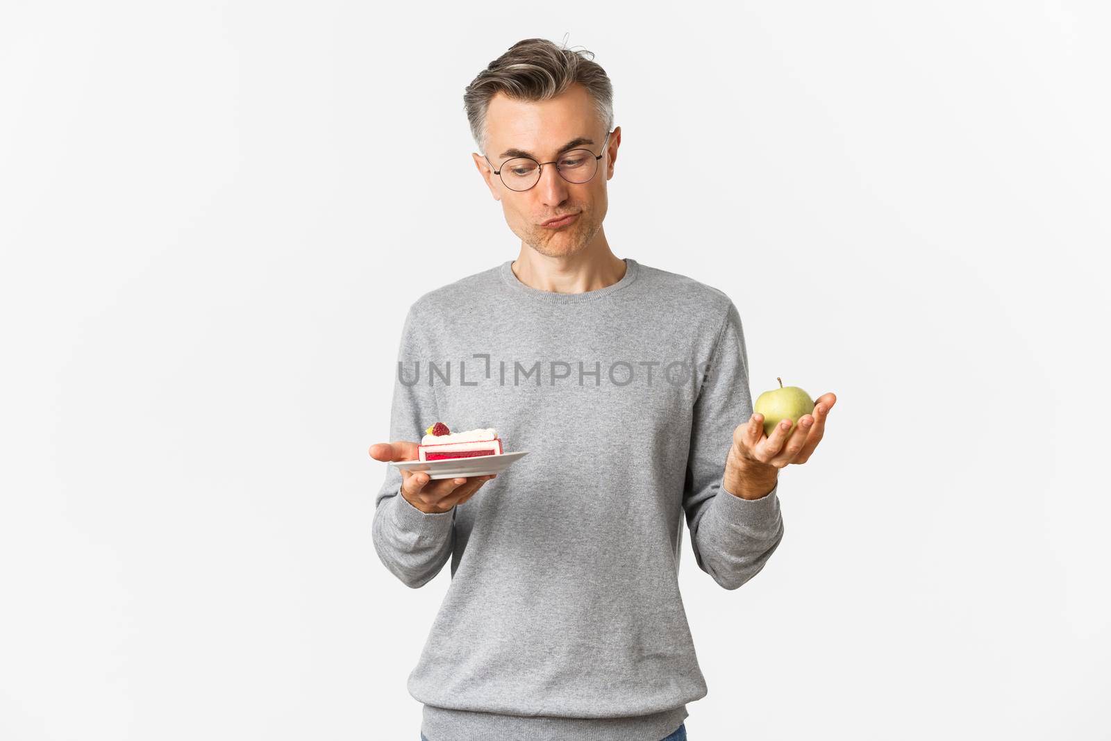 Portrait of thoughtful middle-aged man in glasses, making decision between tasty cake and green apple, standing over white background.