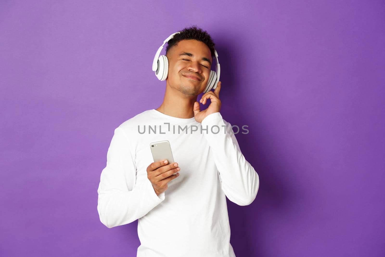 Image of carefree african-american man, enjoying listening music in headphones, holding smartphone and smiling pleased, standing over purple background.