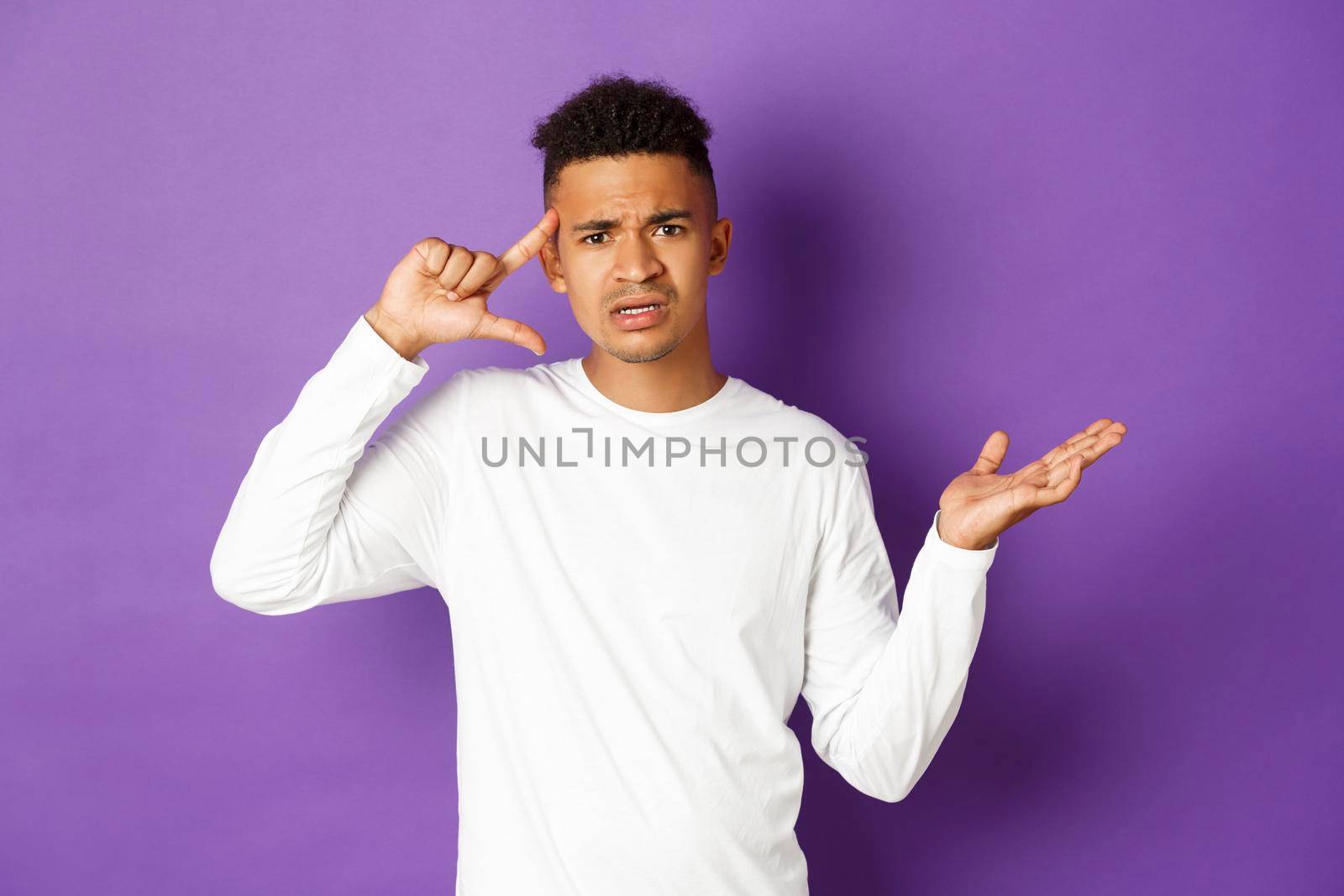 Image of angry african-american man, looking confused, pointing at head and scolding person for stupid action, standing over purple background.