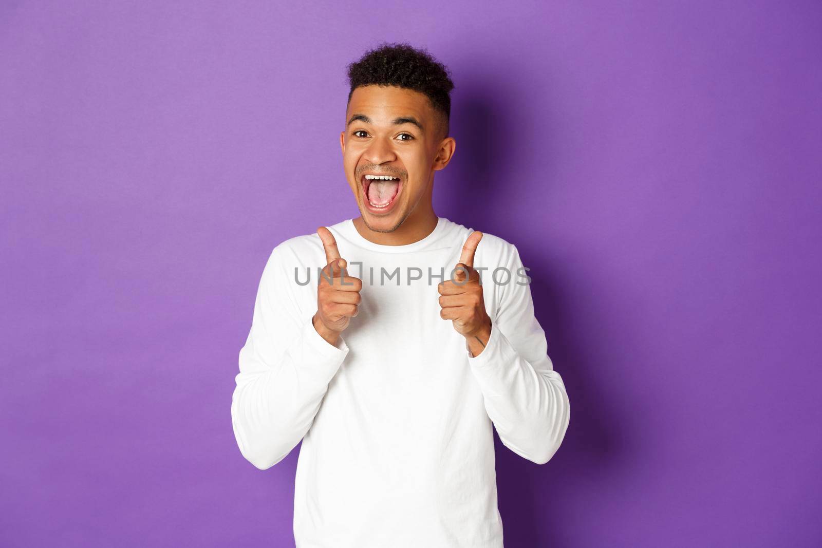 Image of cheerful african-american young guy, pointing fingers at camera and smiling, praise good choice, congratulate with something, standing over purple background.