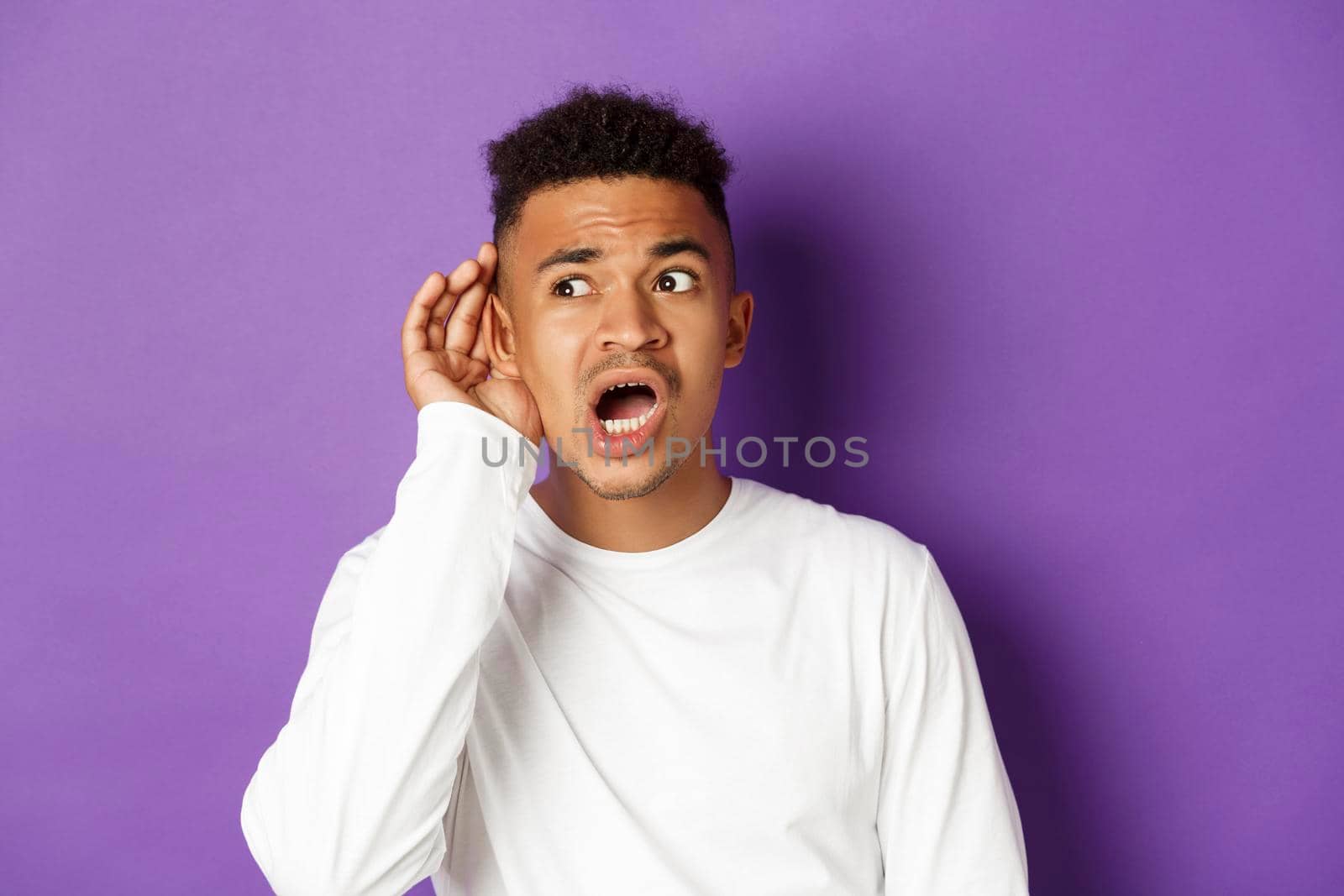 Image of african-american man in white sweatshirt eavesdropping, looking worried, trying to overhead conversation, standing over purple background.