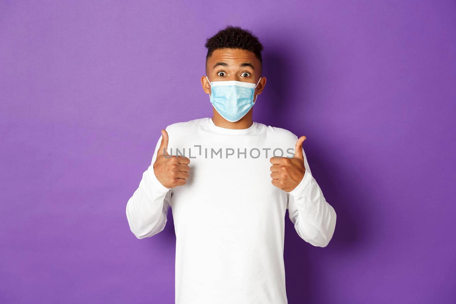 Concept of covid-19, pandemic and social distancing. Amazed african-american guy in medical mask, showing thumbs-up in approval, like something, standing over purple background by Benzoix