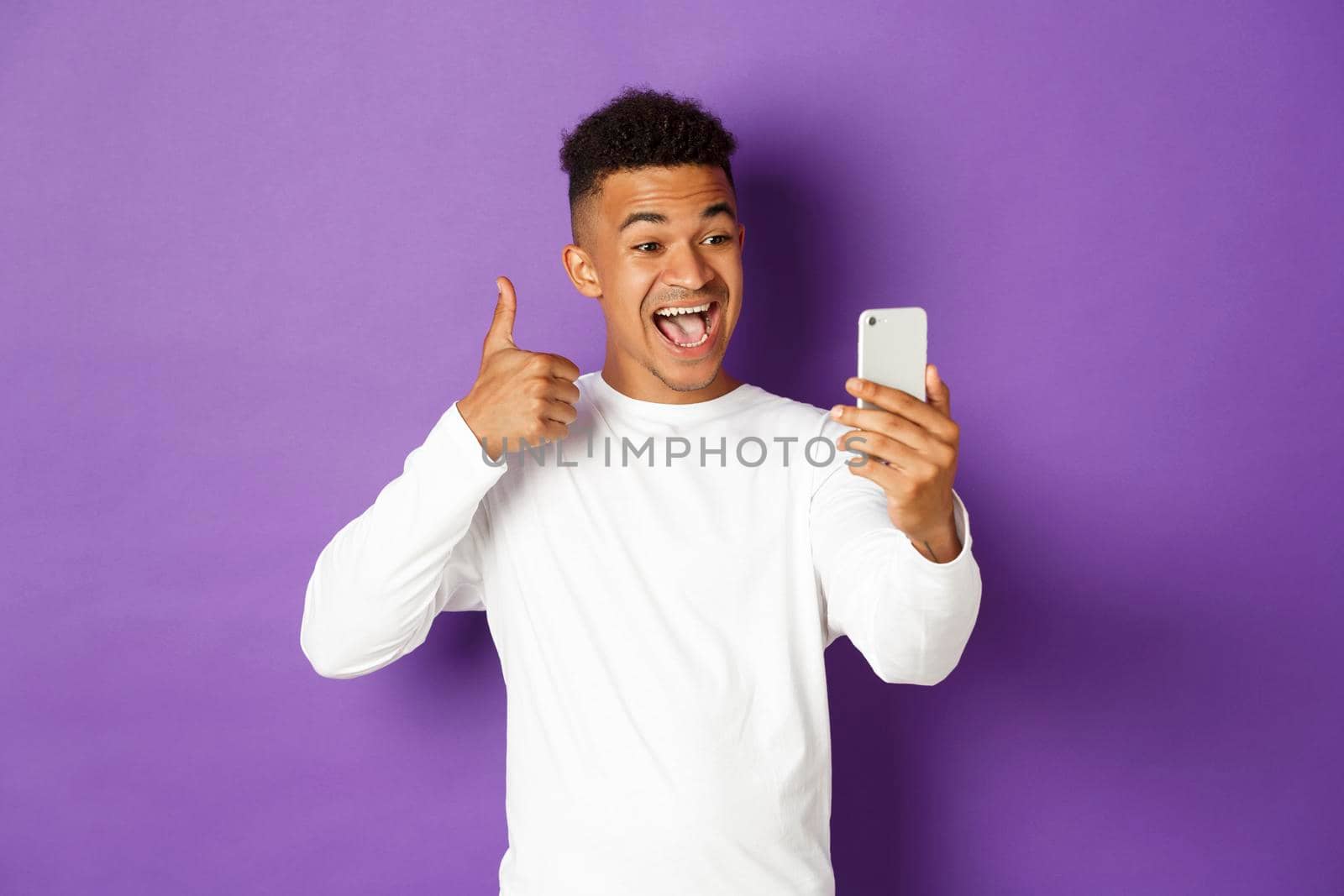 Image of smiling african-american male blogger, showing thumbs-up at mobile phone camera, recommend something, standing with smartphone over purple background by Benzoix