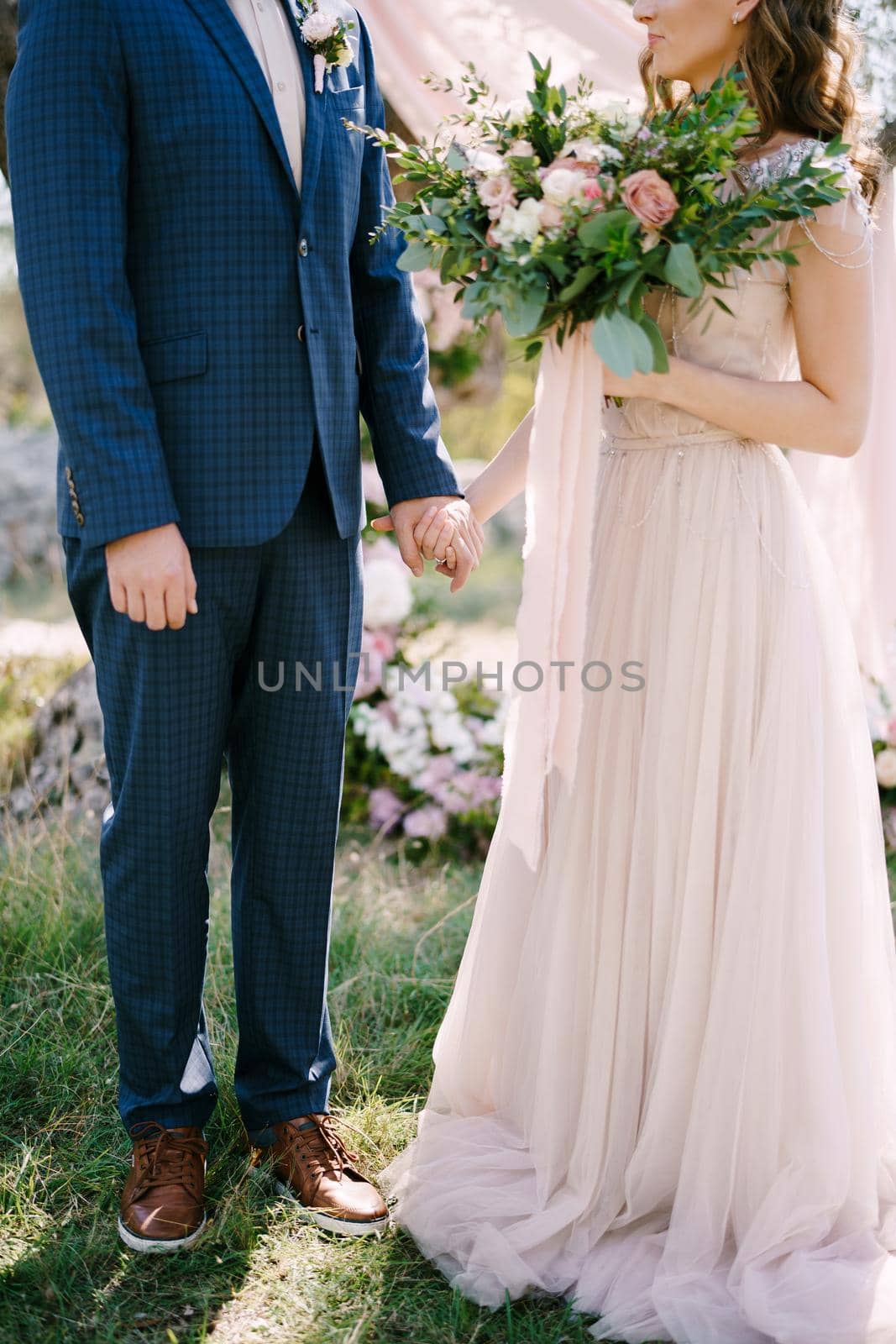 Bride in a long dress with a bouquet stands by the hand with groom in a blue plaid suit. High quality photo