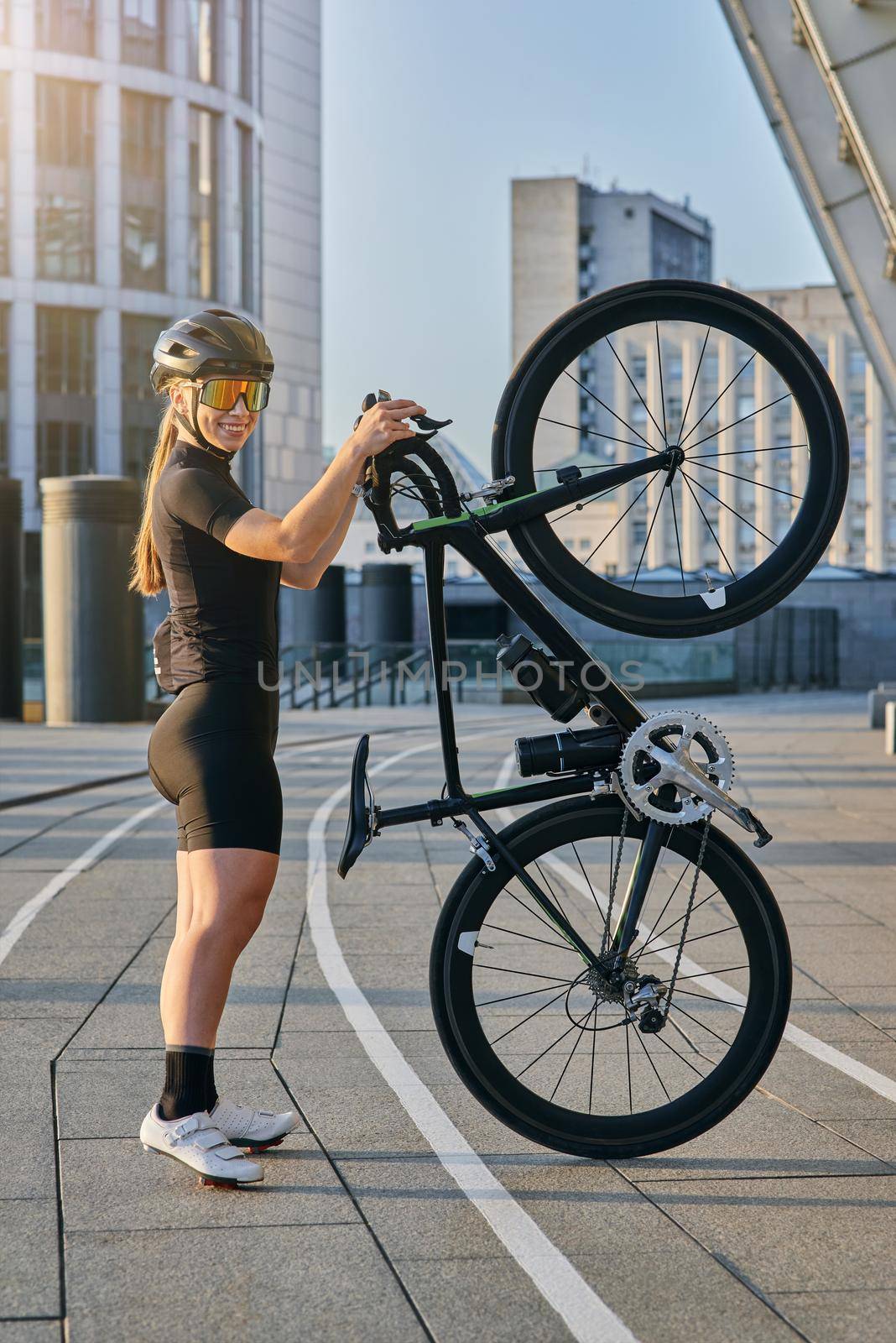 Side view of professional female cyclist in black cycling garment and protective gear smiling at camera, posing with her bicycle standing upright outdoors on a sunny day. Sports concept