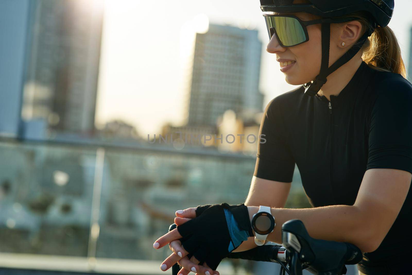 Close up portrait of joyful young sportswoman in black cycling garment and protective gear having a rest, admiring cityscape while training outdoors on a sunny day. Active lifestyle, sports concept