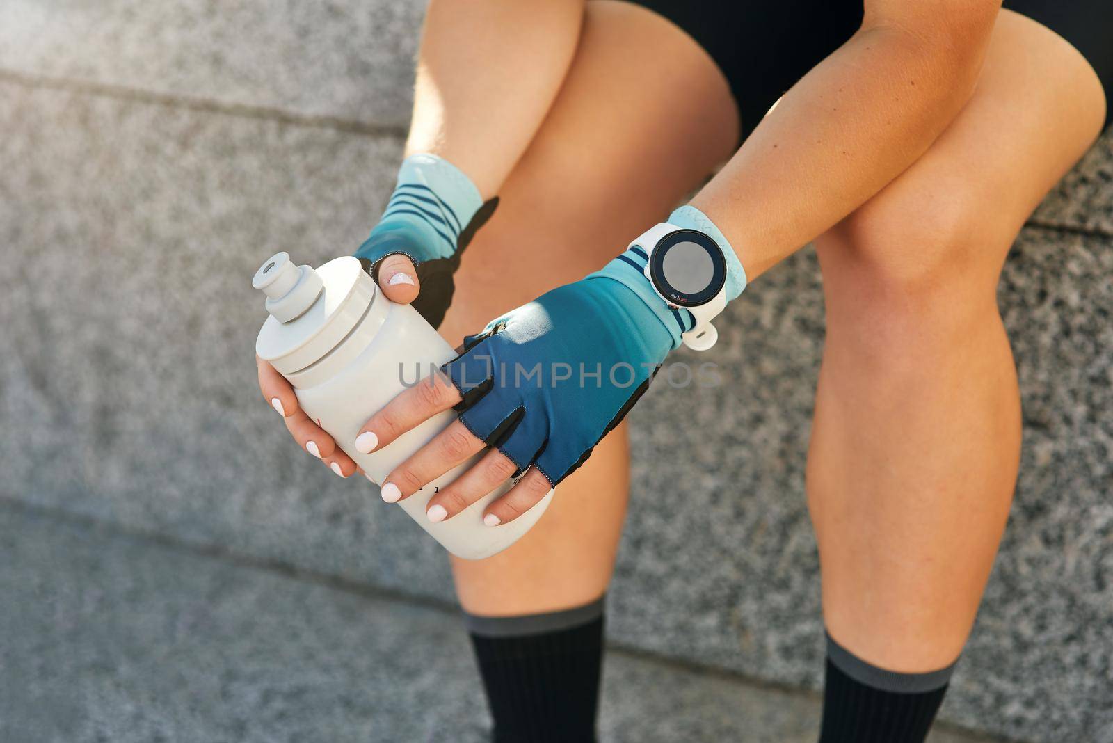 Close up shot of hands of professional female cyclist wearing protective cycling gloves and smartwatch holding drink bottle while sitting, resting after training by friendsstock